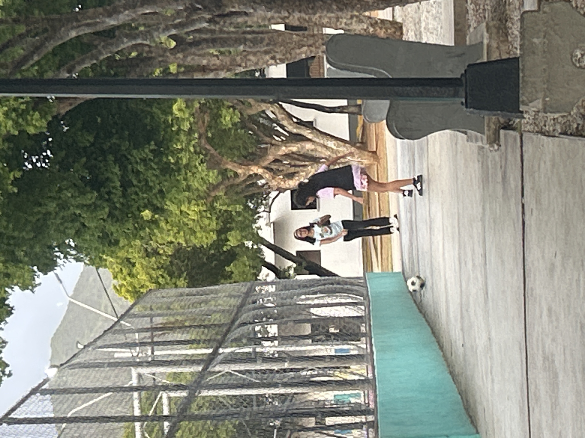 The image depicts an outdoor scene in Santa Ana, Venezuela, likely near a recreational facility. In the foreground, a concrete walkway is visible, bordered by a turquoise retaining wall on the left. A soccer ball rests on the walkway. Two young females are present. One, wearing glasses, a white t-shirt with a graphic, and black pants, stands to the left, holding what appears to be a brown bag. The second, to the right, is in motion, wearing a pink t-shirt, a dark vest, and a patterned skirt. She is actively playing with the soccer ball. Behind them, a chain-link fence encloses a court area, and beyond that, trees and buildings are visible. A tall pole with a light fixture stands on the right side of the walkway. In the background, hills and mountains can be seen under a cloudy sky.