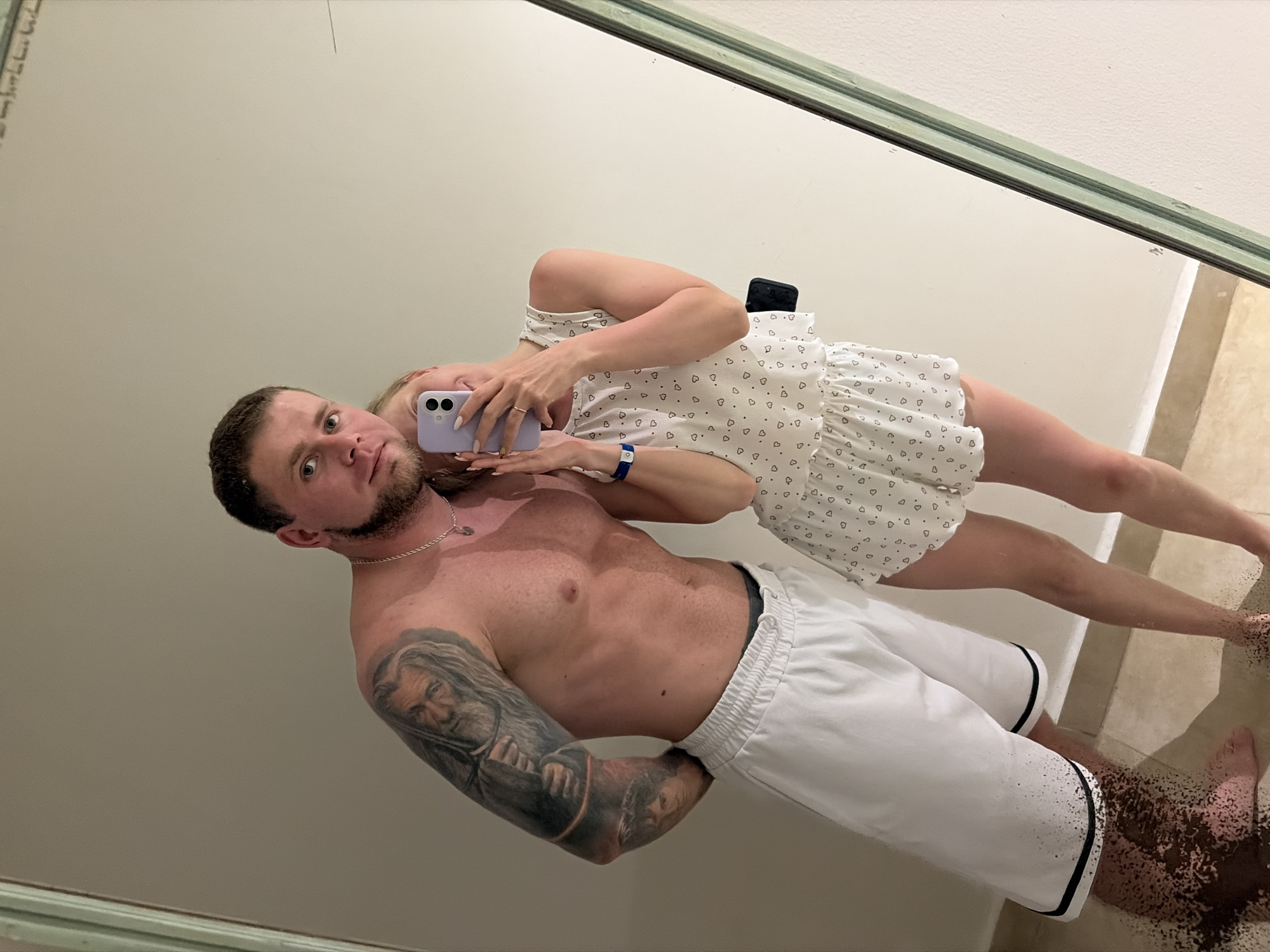 A man and a woman are posing in a selfie in front of a mirror. The man is bare-chested with white shorts, while the woman wears a white dress with a heart pattern. The man has a tattoo on his left arm. The woman holds a purple phone to take the picture. The location appears to be in Sharm El Sheikh, Egypt, based on visual cues. The environment suggests an interior space, possibly a hotel room.