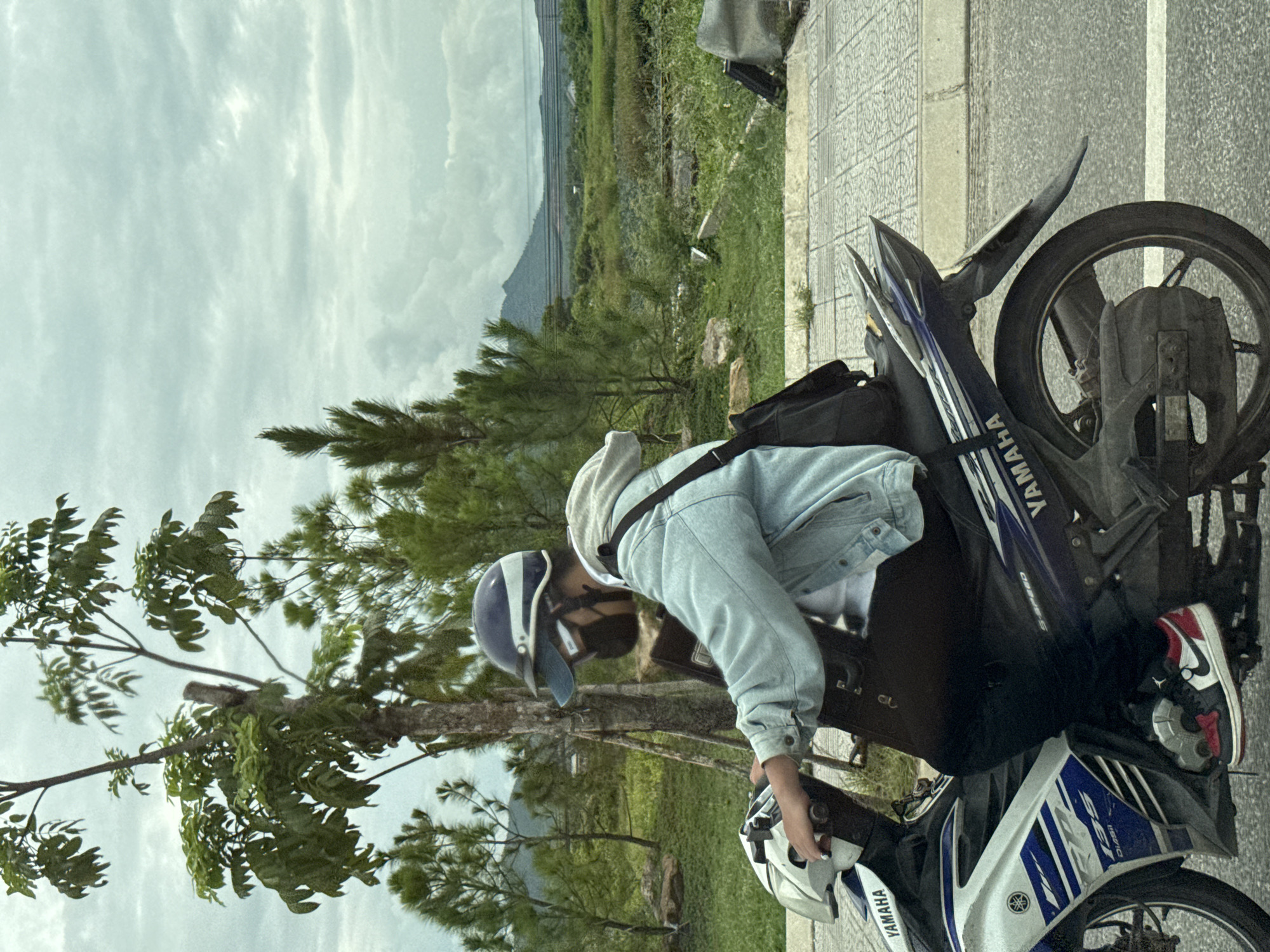 A person wearing a light blue denim jacket, a dark mask, and a blue and white helmet rides a white and blue Yamaha motorcycle. The person is in profile, facing right. The motorcycle has the "YAMAHA" logo visible on its side. The background shows a tree with sparse green leaves on the left, a cloudy sky, and a distant green landscape with a mountain peak. The motorcycle is on a paved road with a tiled sidewalk to the right. The scene appears to be in Kim Trà Ward, Vietnam.