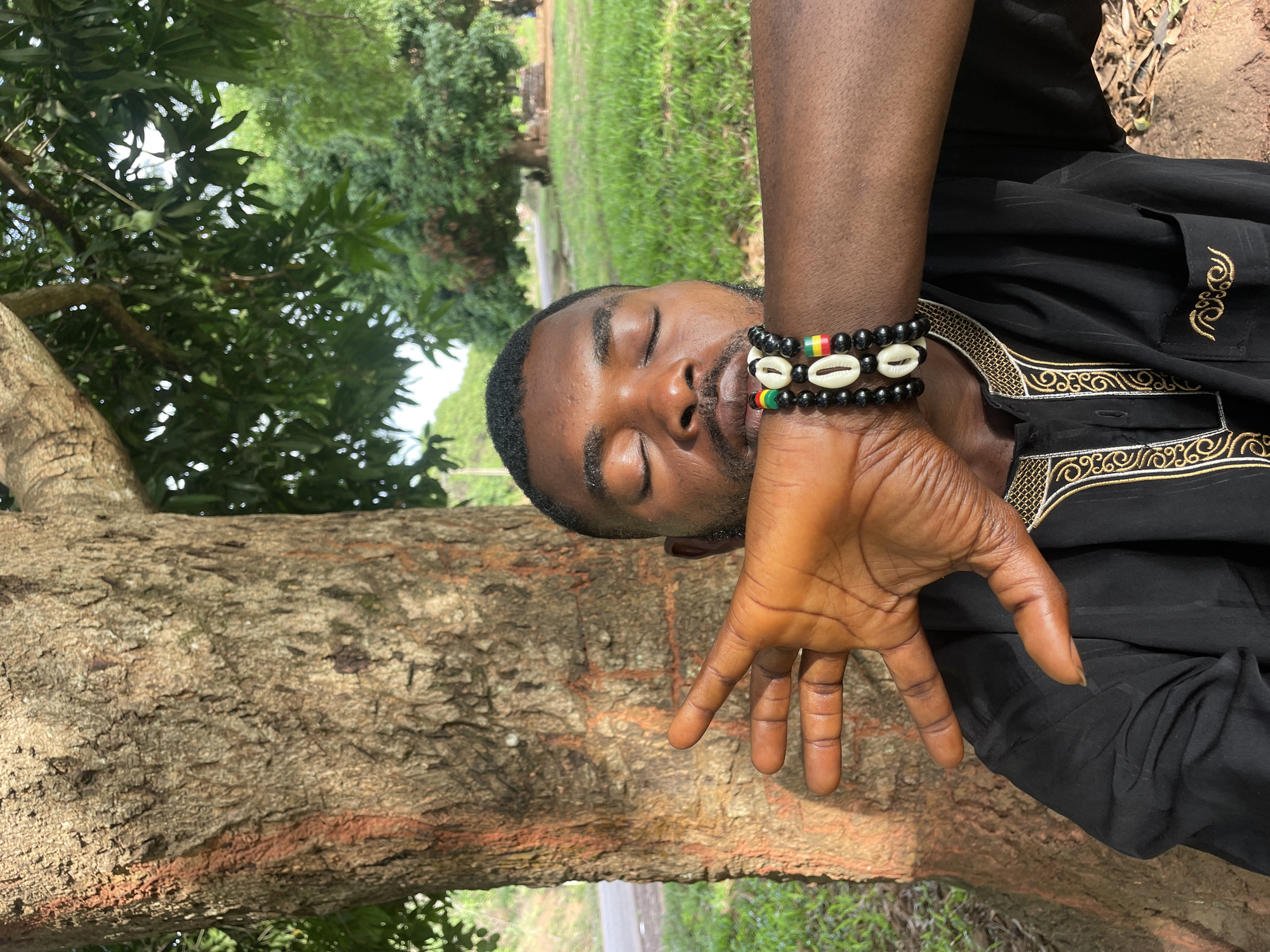 The image shows a man resting in a natural environment, potentially in Ogbe, Nigeria. The man is wearing a black garment with gold embroidery on the collar and sleeves. He is also wearing three bracelets on his left wrist. The bracelets appear to be made of beads and cowrie shells with red, yellow, and green colors on one. He is positioned near a large tree, with his hand extending over the trunk. The background includes a grassy area and other trees, suggesting an outdoor setting. The man's eyes are closed, indicating he is resting or sleeping. The overall impression is one of tranquility and connection with nature.