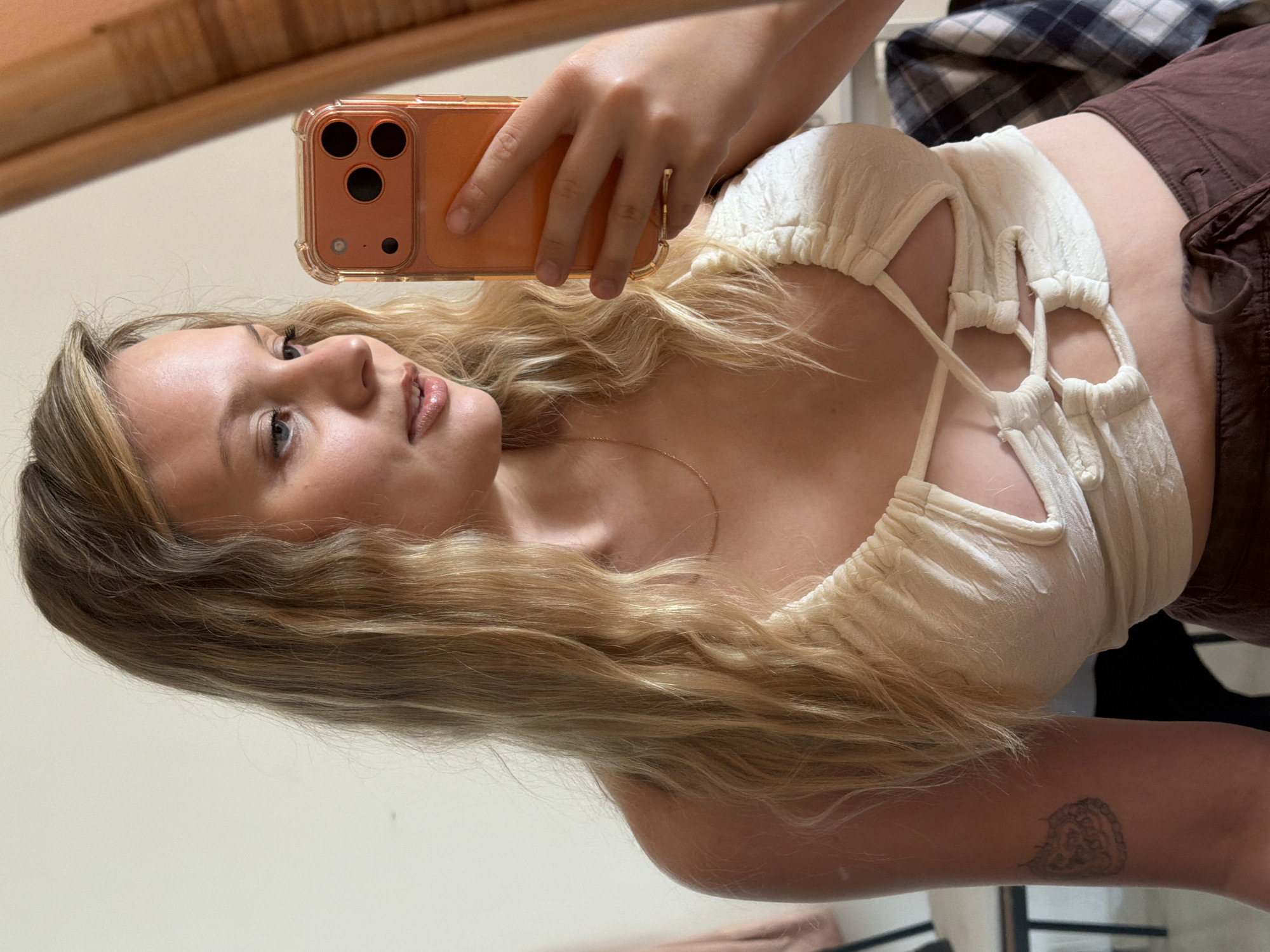 A mirror selfie captures a young adult female indoors. She has long, wavy blonde hair and light blue eyes, looking towards her reflected image. A detailed, possibly floral or mandala-like tattoo is visible on her upper left arm. She wears a cream-colored, textured crop top with frontal cutouts and intricate lacing, along with dark brown drawstring trousers. A thin gold chain necklace adorns her neck, and a gold ring is present on her left ring finger. In her right hand, she holds an orange-cased smartphone, identified as an iPhone by its triple camera array, positioned to capture the reflection. The background features a plain light-colored wall, a dark-framed chair on the left, and a dark plaid fabric draped over an unseen object on the lower right. A vertical wooden strip is partially visible on the far right edge of the frame. The specific location in Tel-Aviv, Israel, cannot be ascertained from the visual information provided.
