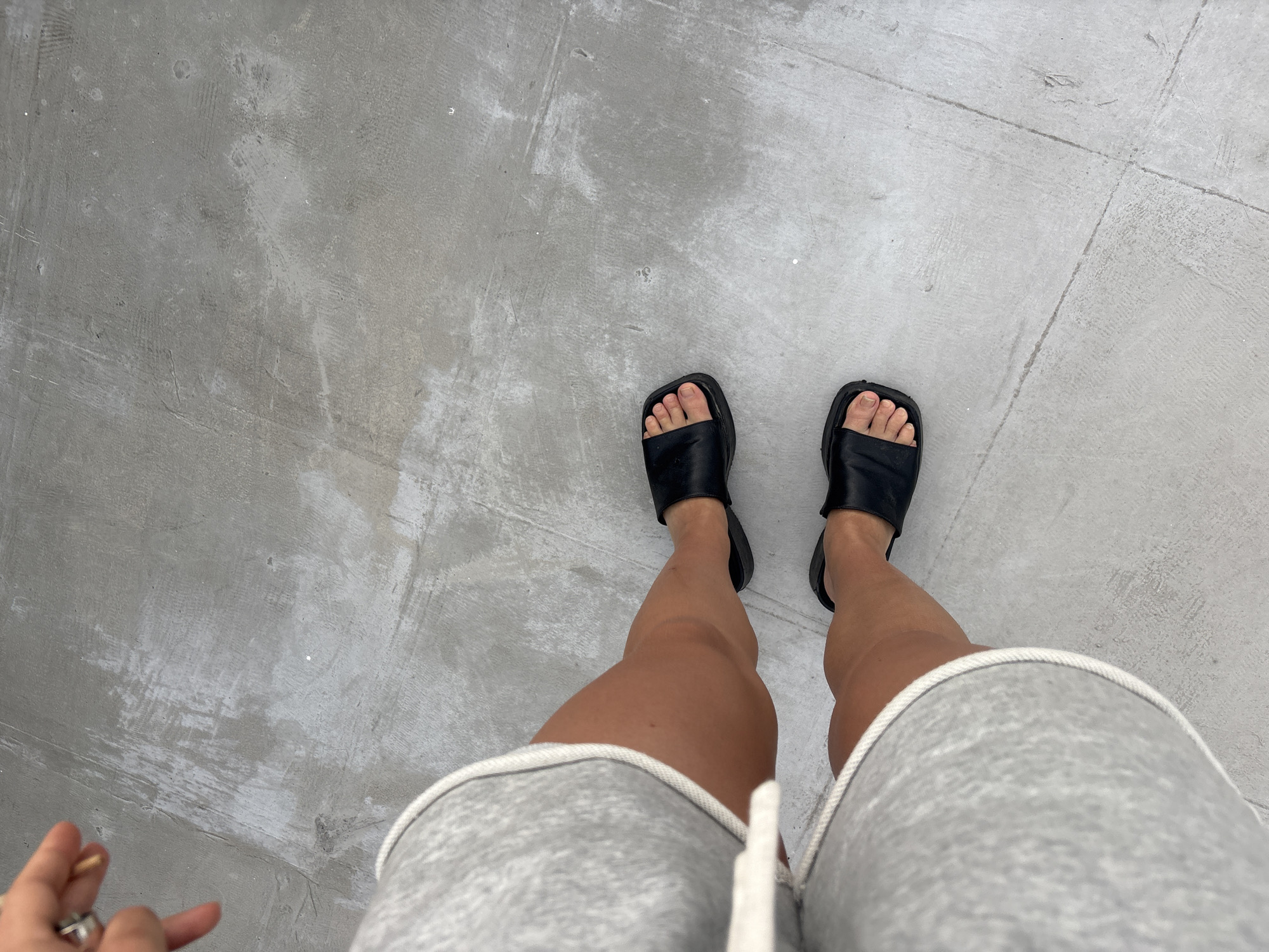 A photograph taken from a first-person perspective shows a person standing on a concrete surface. The person is wearing black leather-like slides and gray sweat shorts with a white drawstring. The concrete surface has a mottled texture and appears to be composed of large square tiles. The person's right hand is partially visible in the lower left corner of the frame. Đà Nẵng, Vietnam, cannot be determined from the image.