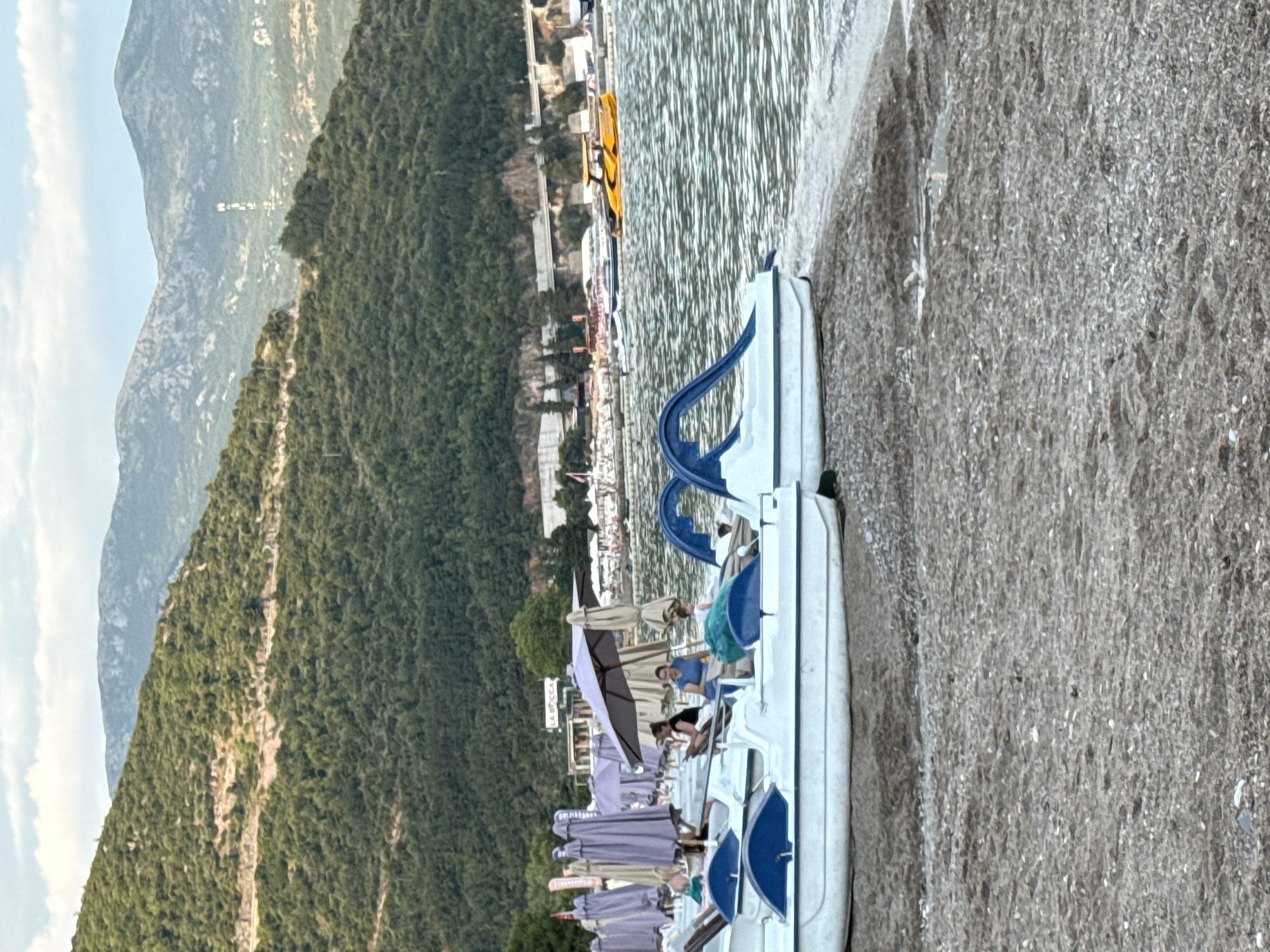 The image depicts a serene scene on a beach in Budva, Montenegro, with the turquoise waters of the Adriatic Sea stretching towards a backdrop of lush green mountains. In the foreground, several pedal boats are neatly lined up on the sandy shore, some partially submerged in the shallow water. A few individuals are seen relaxing in the pedal boats, enjoying the gentle sea breeze. Umbrellas are scattered across the beach, providing shade to beachgoers who are either seated or strolling along the coastline. In the distance, a yellow speed boat is navigating the waters, adding a touch of vibrancy to the scene. The mountains in the background are covered with dense vegetation, with a few buildings and a road visible along their slopes, hinting at the developed landscape of Budva. The sky above is a soft blue, with scattered clouds, creating a tranquil and picturesque atmosphere. The image captures the essence of a perfect summer day at the beach, offering a glimpse into the relaxed lifestyle and natural beauty of Budva, Montenegro.