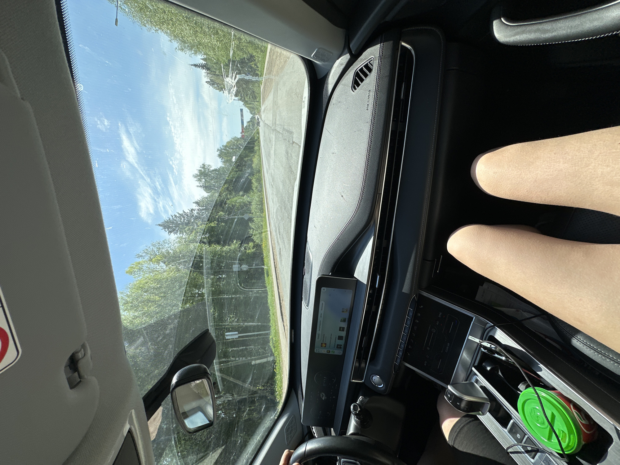 The image depicts the interior of a car, viewed from the driver's seat. The dashboard is visible, featuring a digital display screen and various control panels. A drink with a green lid is present in the center console. A person's leg is visible on the passenger side. Through the windshield, a road is seen with trees lining the sides. The sky is partly cloudy. The specific location within the city of Uva, Russia cannot be determined from the image.