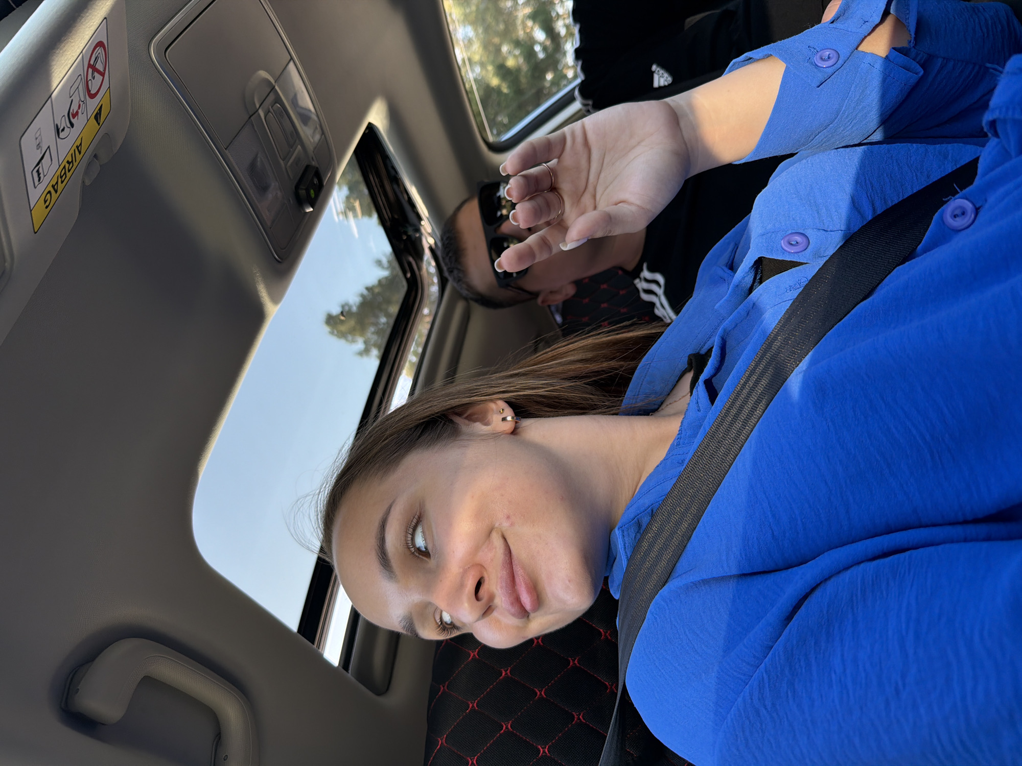 A woman wearing a blue shirt and a seatbelt is in a car, posing for a picture with a playful expression. Behind her, another person is wearing sunglasses and a mask. The car has a sunroof, allowing a view of trees and the sky outside. The image was taken in the interior of a car, in daylight, and is slightly rotated. The location is not detectable from the information in this image.