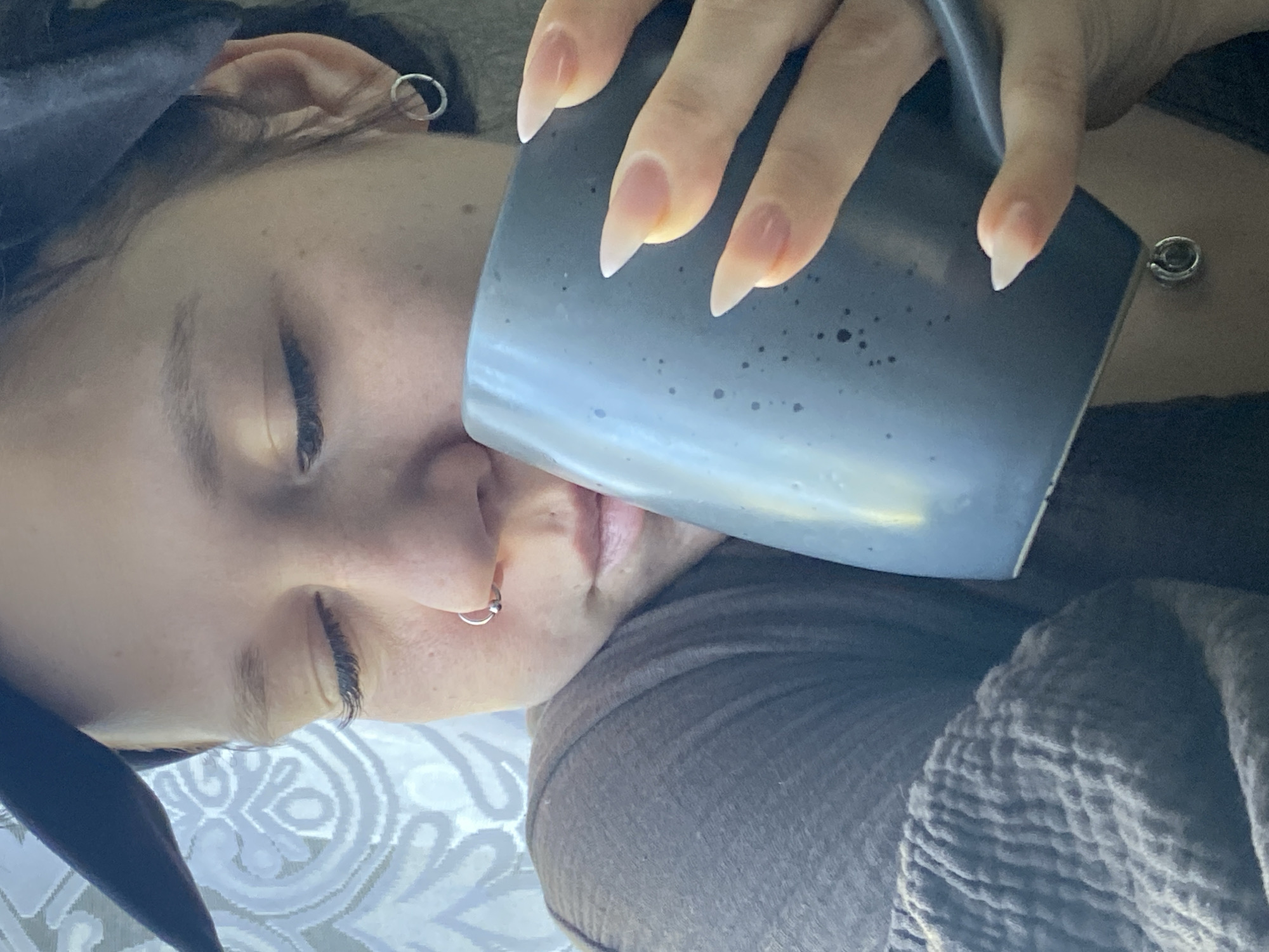 A person lies in bed, face tilted toward a gray-blue mug held by their hand with long, pointed nails. The person has closed eyes, a nose ring, and an earring in their left ear. They are wearing a dark headband and are covered by a brown blanket on a patterned bedsheet. The location is not discernible in the image and cannot be confirmed as Chaikovsky, Russia.