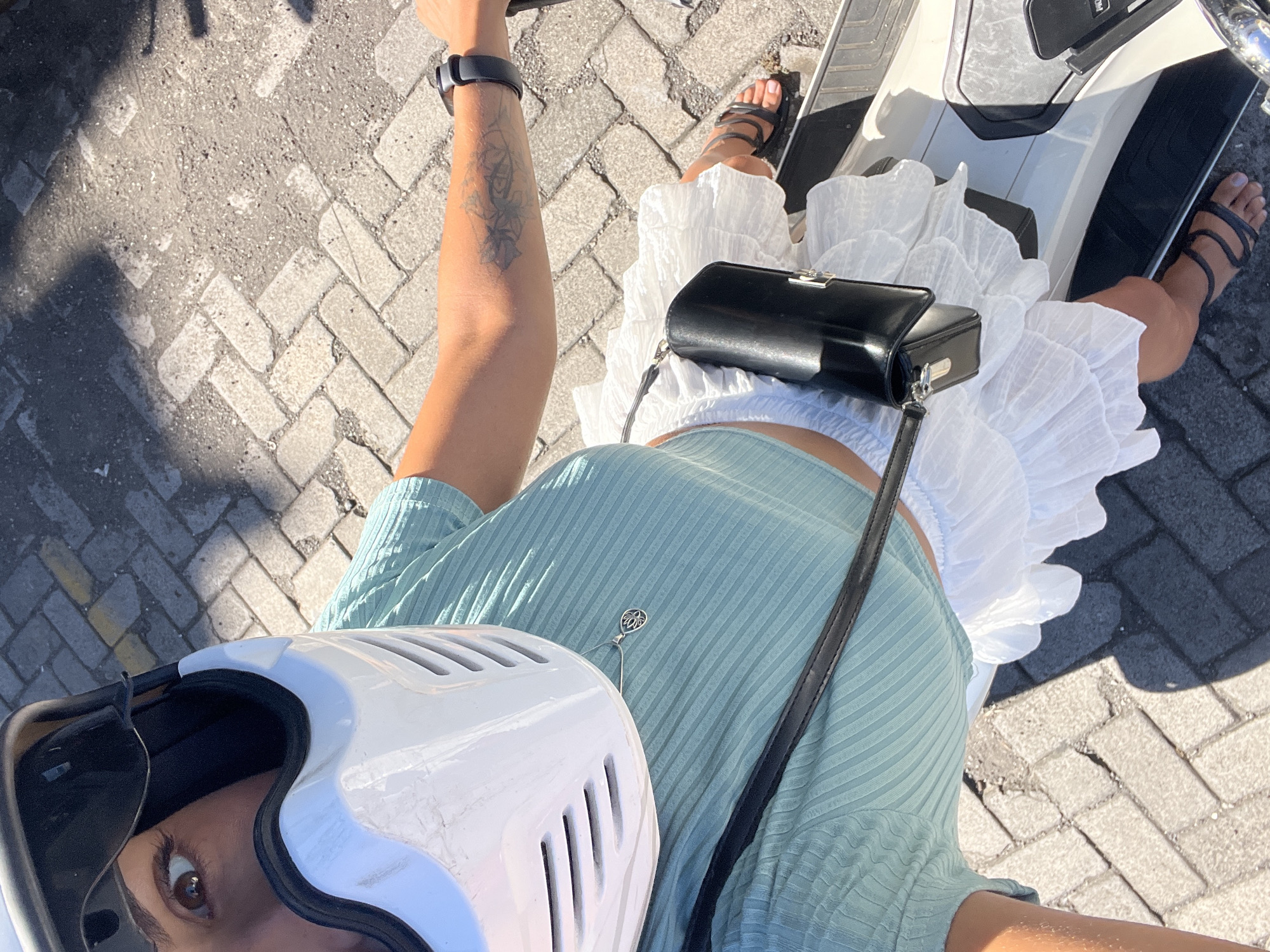 A high-angle shot captures a woman seated on a white moped, likely within the city of Kuta Selatan, Indonesia, as suggested by the local street pavement. The woman wears a light green, short-sleeved ribbed shirt, a white ruffled skirt, and strappy black sandals. She sports a black smartwatch on her left wrist and has a tattoo on her forearm. A small black purse hangs across her body, its strap extending down her side. She is wearing a white helmet with a black visor, obscuring much of her face except for one visible eye. The moped's white body is partially visible beneath her. The paved street surface, constructed of gray interlocking bricks, is evident in the background. Sunlight casts shadows across the scene.