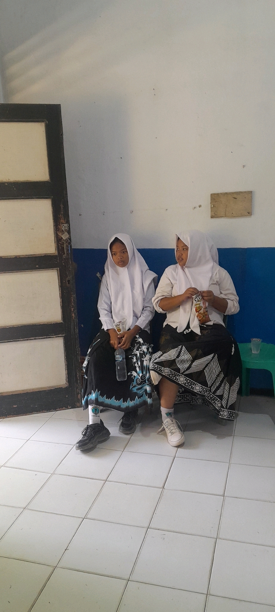Two young females are sitting on a bench in front of a wall. Both are wearing white headscarves and long black skirts with white patterns. The female on the left is holding a bottle of water and the female on the right is holding a snack bag. The floor is tiled and there is a door on the left side of the image. The wall behind them is painted white and blue. A small green stool is visible on the right side of the image. The location is identified as Tahunan, Indonesia.