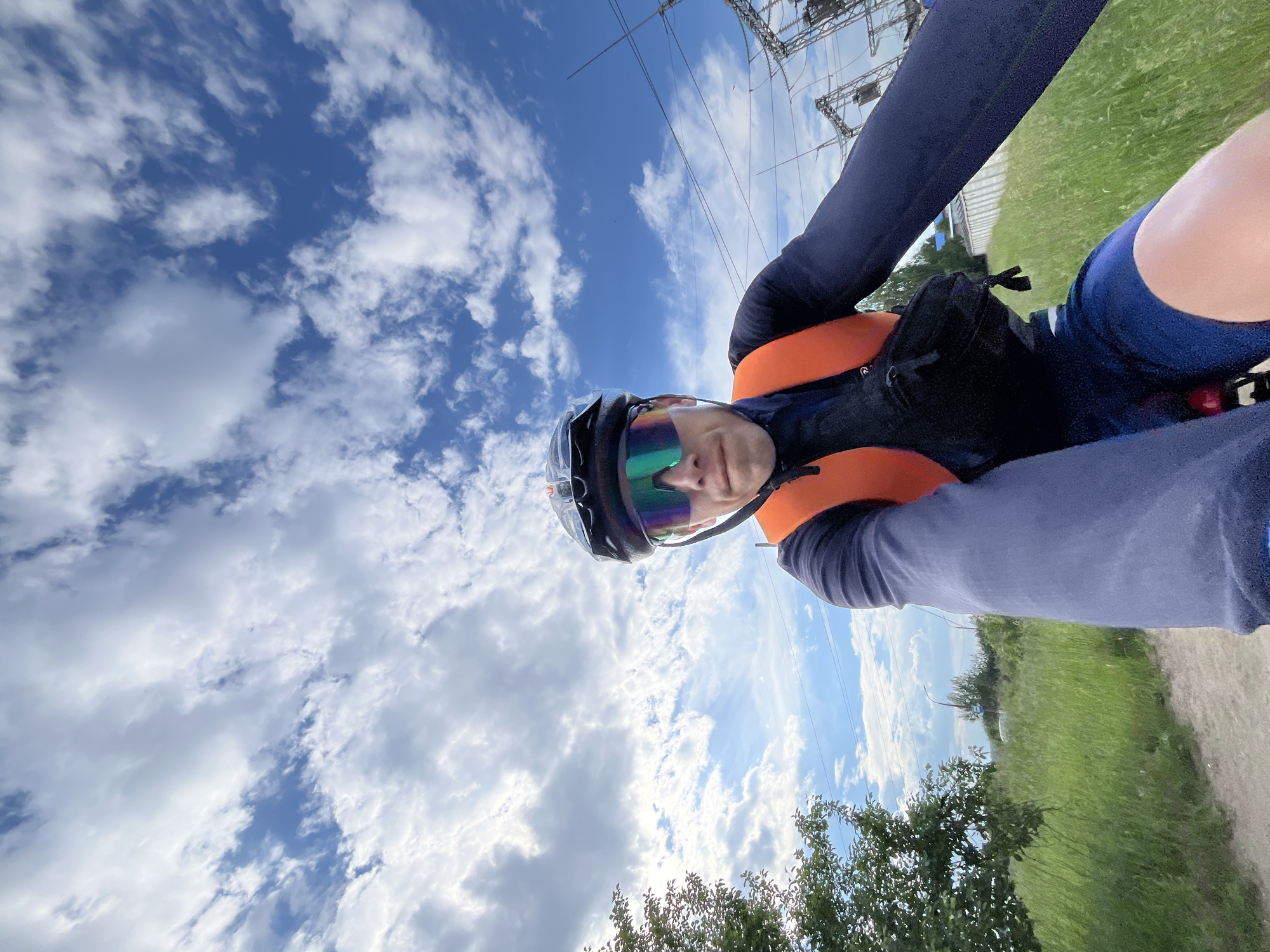 A close-up, selfie-style, eye-level shot shows a male cyclist in Chaikovsky, Russia, on a cloudy day. The cyclist, wearing a helmet, neon-tinted sunglasses, a navy long-sleeved shirt, an orange vest, and navy shorts, faces the camera. A black fanny pack is worn across his chest. The cyclist is in motion, evident from the slight blur in the background.
The environment is a rural or suburban area with a dirt path in the foreground, flanked by tall green grass. To the left, a leafy green tree is visible. In the background, power lines and industrial electrical equipment are prominent, suggesting the presence of a power substation or similar infrastructure. The sky is a vibrant blue with scattered white clouds. The scene depicts an outdoor recreational activity in a mixed industrial and natural setting.