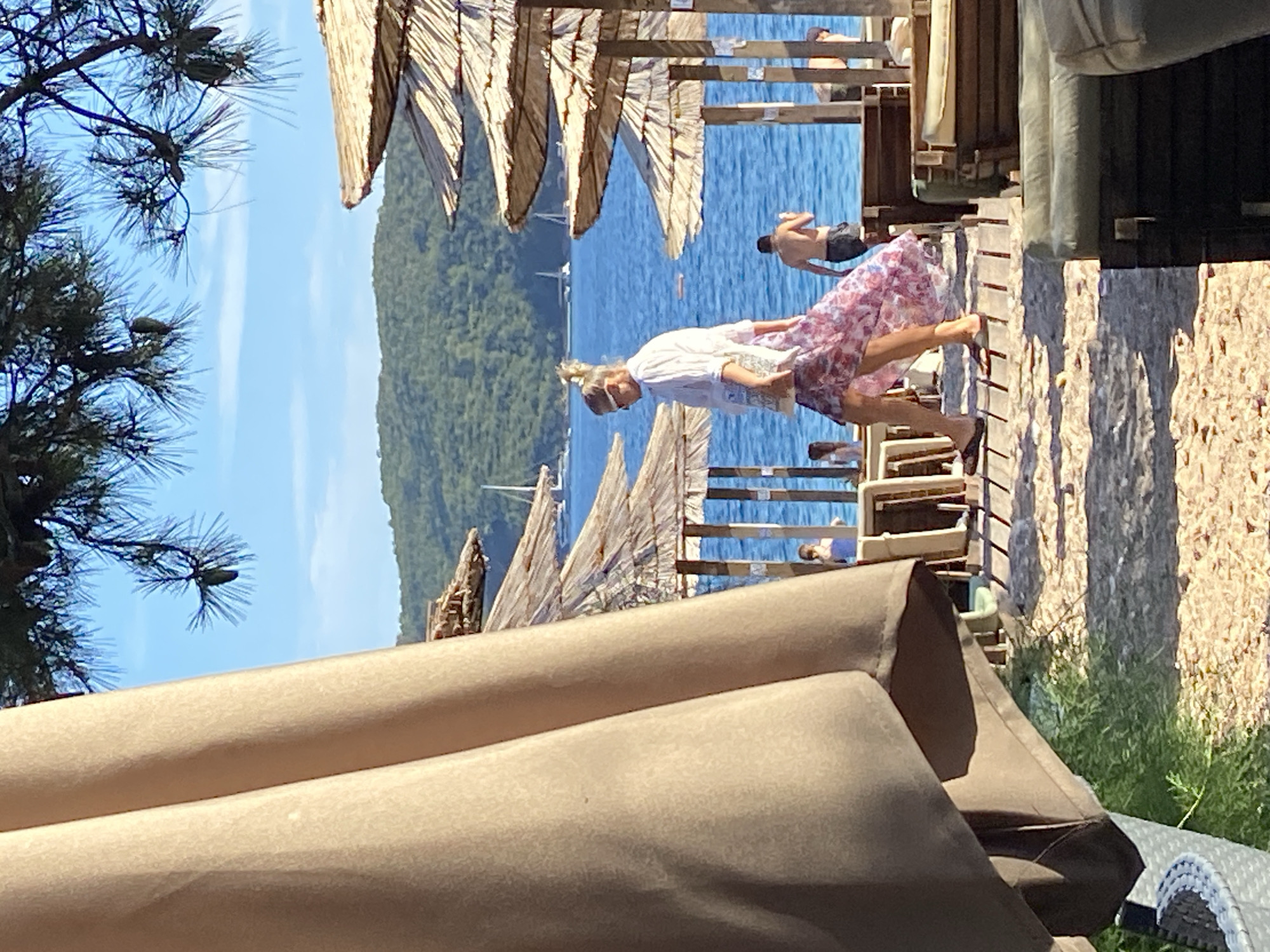 The image depicts a sunny day at a beach resort in Budva, Montenegro, characterized by a clear blue sky and calm sea. In the foreground, a large light-colored umbrella partially obstructs the view of a wooden walkway and thatched-roof beach umbrellas. A woman with blonde hair and sunglasses is walking on the walkway, wearing a white shirt and a long, floral-patterned skirt. She is carrying a tote bag. To her right, a man is standing by the water, facing away from the camera. The background features lush green hills sloping down to the sea, with several sailboats visible on the water. Beach chairs and loungers are arranged on the sand and the walkway, suggesting a leisure environment. The overall scene conveys a relaxed, vacation atmosphere.