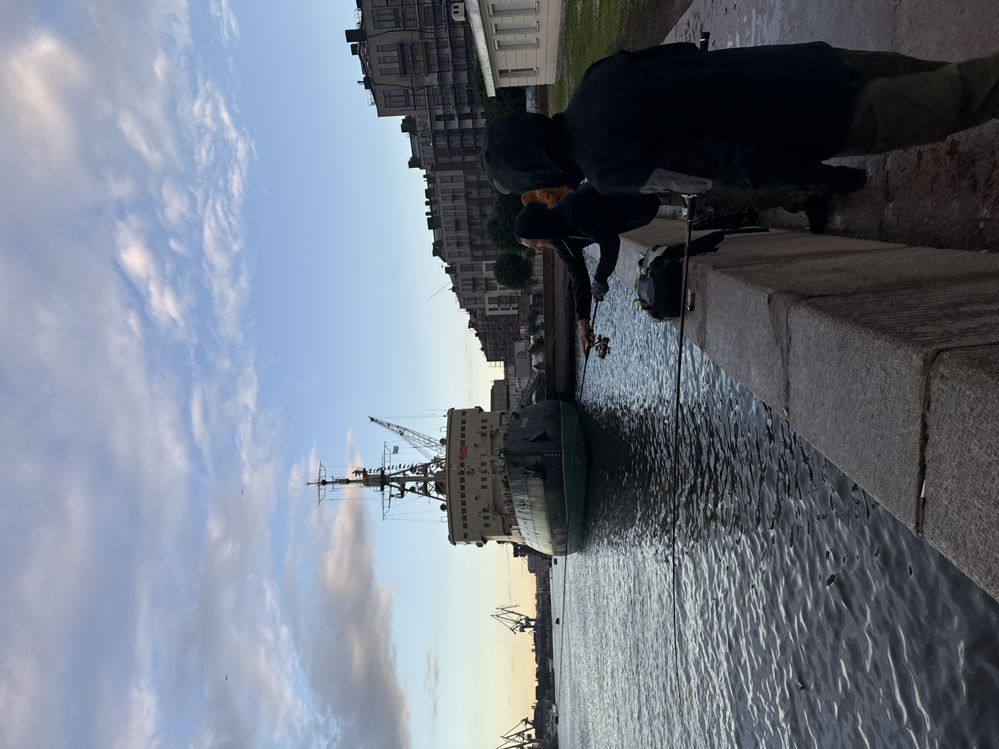 Three individuals are fishing from a stone embankment by a river or canal in Saint Petersburg, Russia, with a large icebreaker ship docked nearby. The icebreaker, named "Krasin" (Красин) as visible on its hull, is dark green and black with a prominent mast and crane. In the background, the Saint Petersburg skyline features apartment buildings, some under construction with scaffolding, and industrial cranes. The sky is partly cloudy with hues of blue and gray, indicating either early morning or late evening light. The water is calm with ripples, reflecting the sky and the icebreaker. The people are dressed in casual, outdoor clothing, and one appears to be actively casting a fishing line. Their focus is on fishing, with fishing rods and a bag visible on the embankment. The scene captures a moment of leisure activity by the waterfront in an urban, industrial setting.