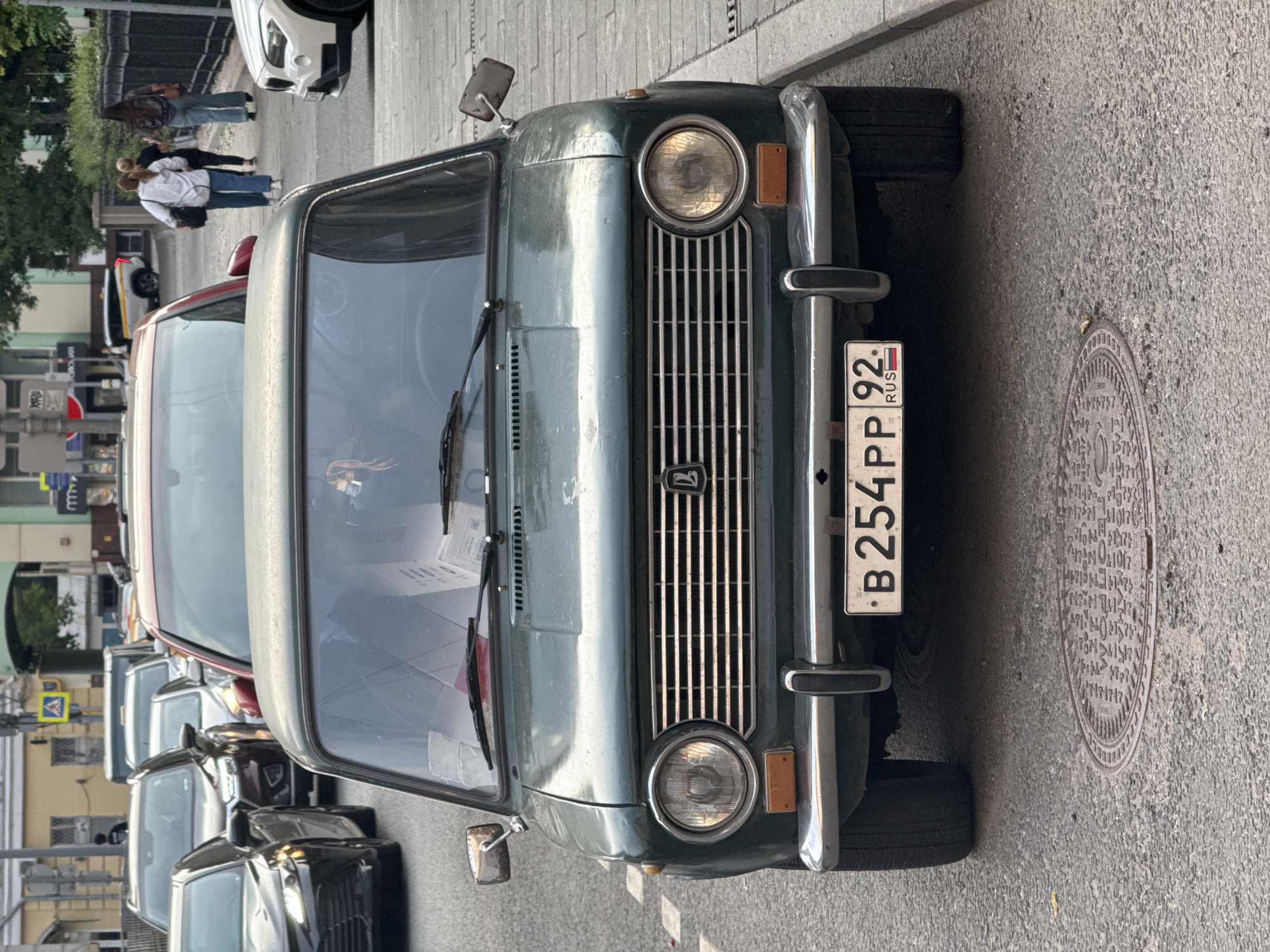 A front view of a dark green, older model Lada car is captured in Moscow, Russia, on a paved street. The car is parked in what appears to be a traffic queue, with other vehicles visible in the background. The license plate of the Lada reads "B 254 PP 92" with a Russian flag emblem. The car's headlights are illuminated, and the grille features horizontal slats with a Lada emblem at its center. A manhole cover is present on the asphalt in front of the car. In the background, a few pedestrians are walking on a sidewalk next to a fence. The environment suggests an urban setting with buildings and traffic signals visible in the distance.