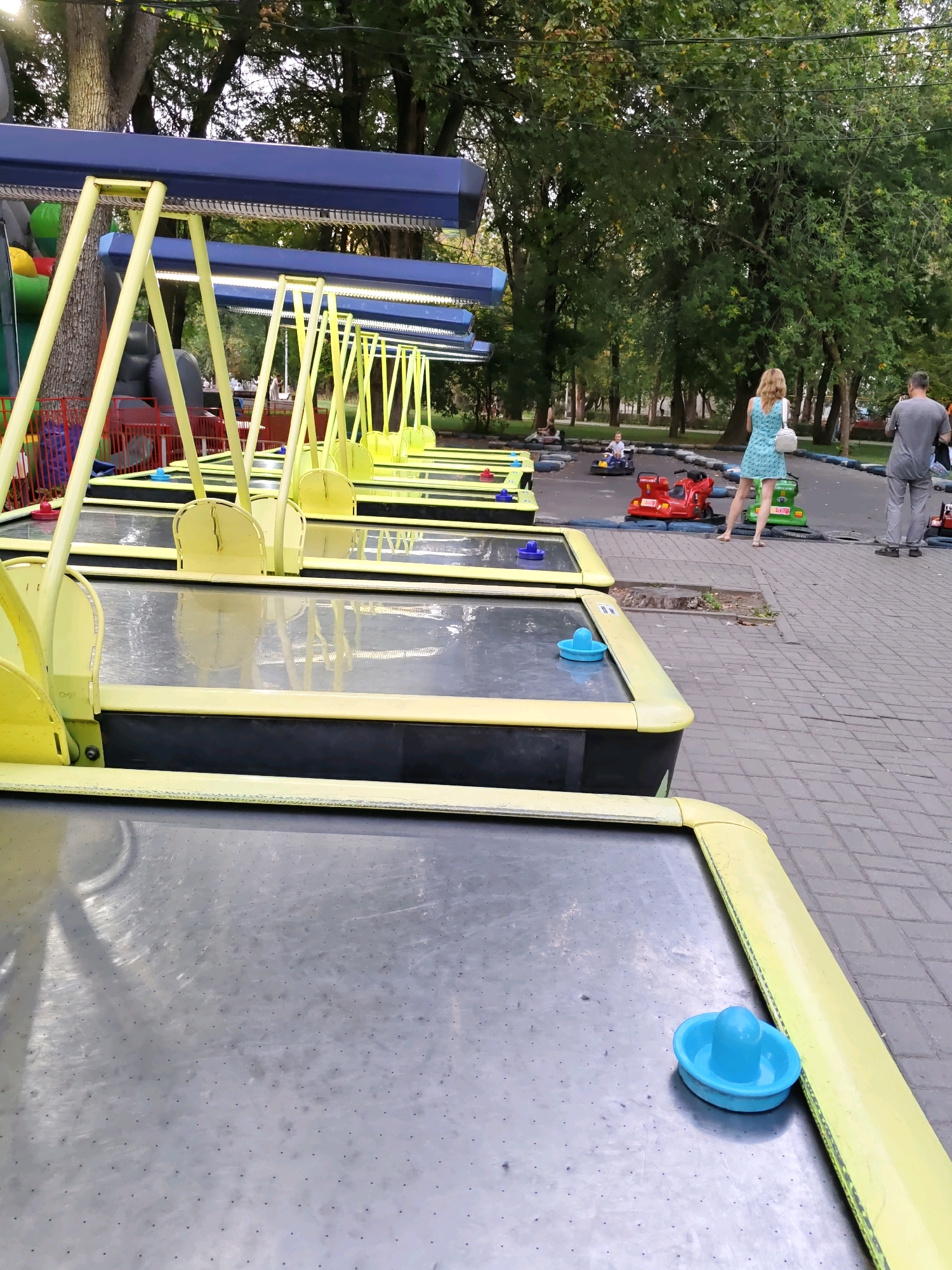 The image depicts an outdoor area that appears to be an amusement park or recreational zone. In the foreground, several air hockey tables are lined up, featuring bright yellow borders and metallic playing surfaces with small air holes. Blue pucks are visible on some of the tables. In the mid-ground, a paved path separates the air hockey tables from a small track with ride-on cars for children. A young child is seated in a small red car on the track. Further in the background, two adults are standing. One is a woman wearing a blue dress and carrying a white bag, facing away from the camera. The other is a man wearing grey pants and a grey shirt, also facing away. The environment is lush with mature trees providing a canopy overhead. The pavement is made of interlocking bricks. The lighting suggests it is daytime, possibly late afternoon. This scene is located in the Rostov-on-Don Botanical Garden in Rostov-on-Don, Russia, specifically within the area designated for children's entertainment.