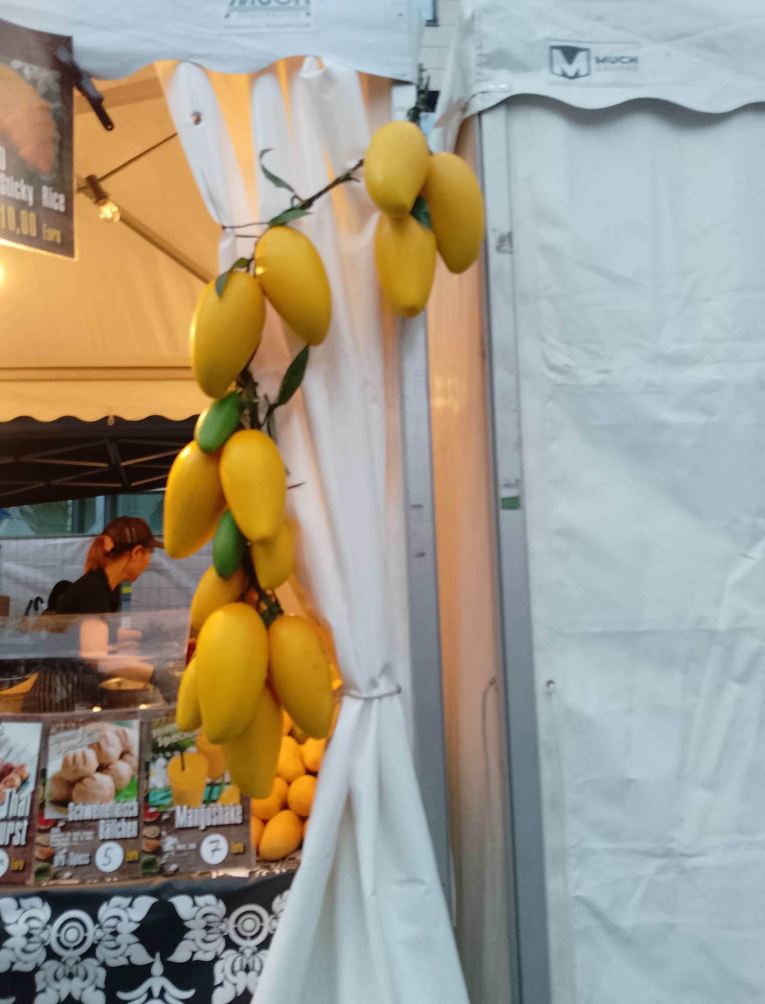 The image shows a street food stall with a display of yellow, mango-shaped objects hanging from a string. A woman with auburn hair pulled back is visible behind a counter, looking to her right. She is wearing a dark top. In front of the stall, there are several signs with text and images of food items, including what appears to be a sausage, bread rolls, and a mango shake. The stall is covered by a white tent. The background shows a white tent and a silver metal pole. The lighting suggests it is daytime or under artificial lighting from the stall.