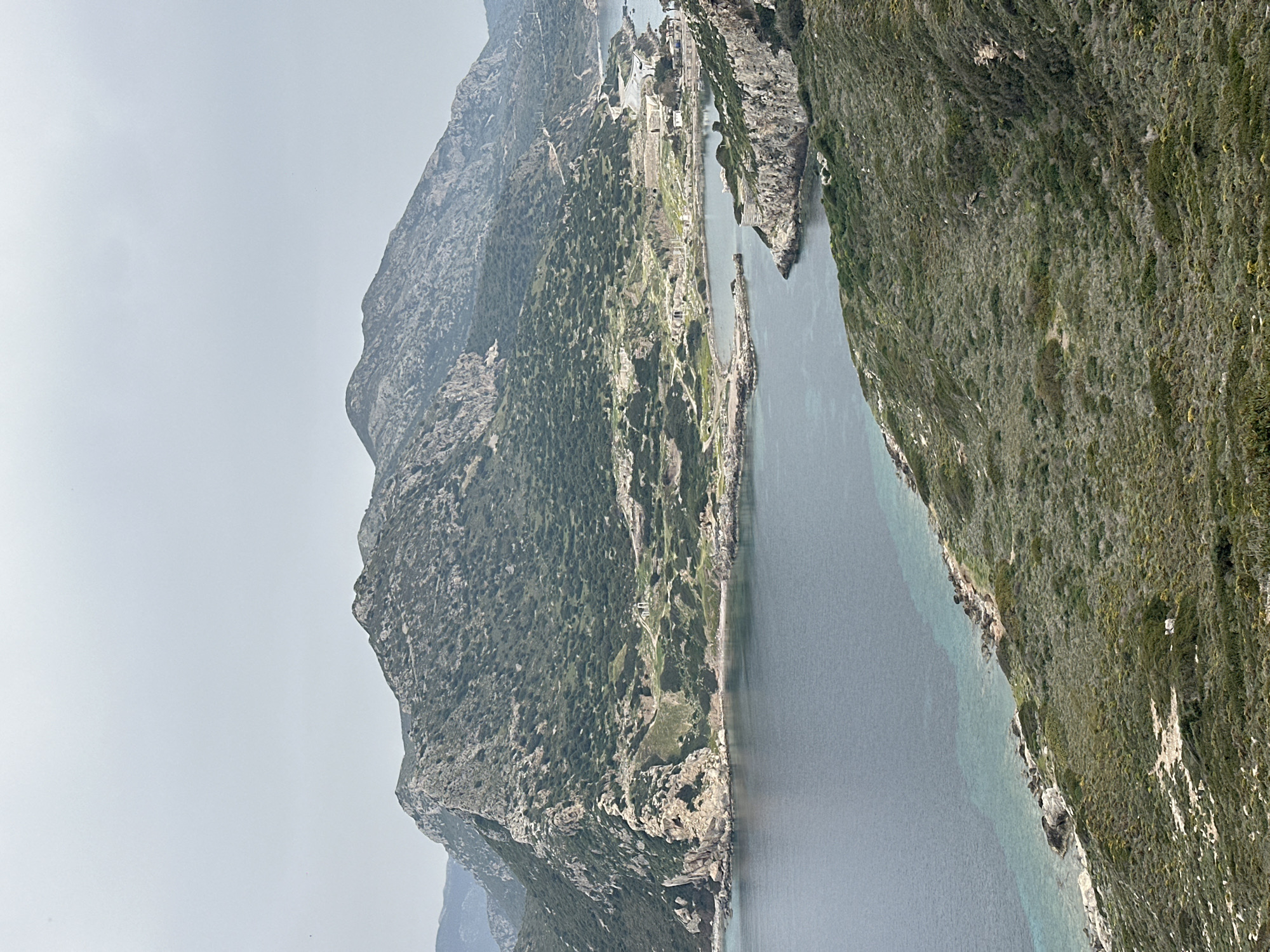 This panoramic image, taken in Datça, Turkey, captures a rugged coastal landscape under a hazy sky. The scene is dominated by a large, steep mountain range stretching across the left side of the frame, from the background towards the midground. Its slopes are densely covered with dark green vegetation, indicating a lush but rocky terrain.
A calm body of water, likely a bay or a strait, occupies the center and right side of the view. The water displays varying shades of blue and gray, with lighter, turquoise tones closer to the shoreline, suggesting shallow areas. Along the base of the left mountain range, a narrow strip of land extends into the water. This strip hosts a small settlement, characterized by a cluster of light-colored buildings and visible roads, possibly including a small port or dock area. Another heavily vegetated hillside, similar in appearance to the distant mountains but closer to the foreground, frames the right side of the image, sloping down towards the water's edge.
The sky is a soft, light gray, indicative of an overcast day or atmospheric haze, which diffuses the sunlight and creates a serene, somewhat muted light across the landscape. There are no people or specific activities visible, emphasizing the natural beauty and tranquility of the scene. No discernible text is present in the image. The time of day appears to be daytime, with soft lighting.