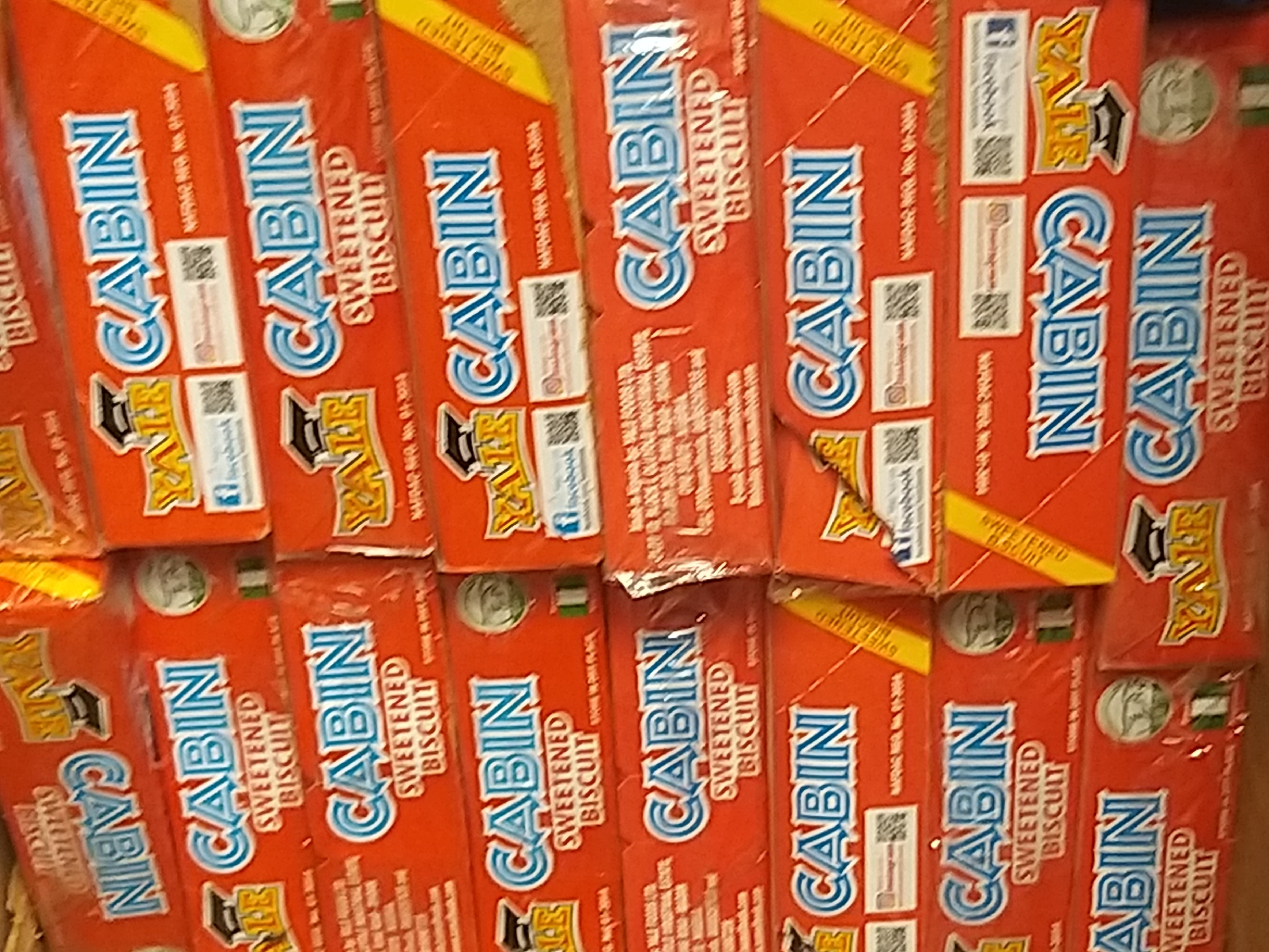 The image shows a stack of red boxes of "CABIN SWEETENED BISCUIT" and some "YALE" branded items. The boxes are stacked in a store or market setting, likely in Kano, Nigeria, given the "Nafdac Reg. No." which is a Nigerian regulatory agency. The boxes display the brand name "CABIN" in blue and white lettering, with the words "SWEETENED BISCUIT" below. Some boxes also feature QR codes and social media icons, suggesting they are modern consumer products. The lighting is bright, indicating an indoor or shaded outdoor environment. No people are visible in the image.