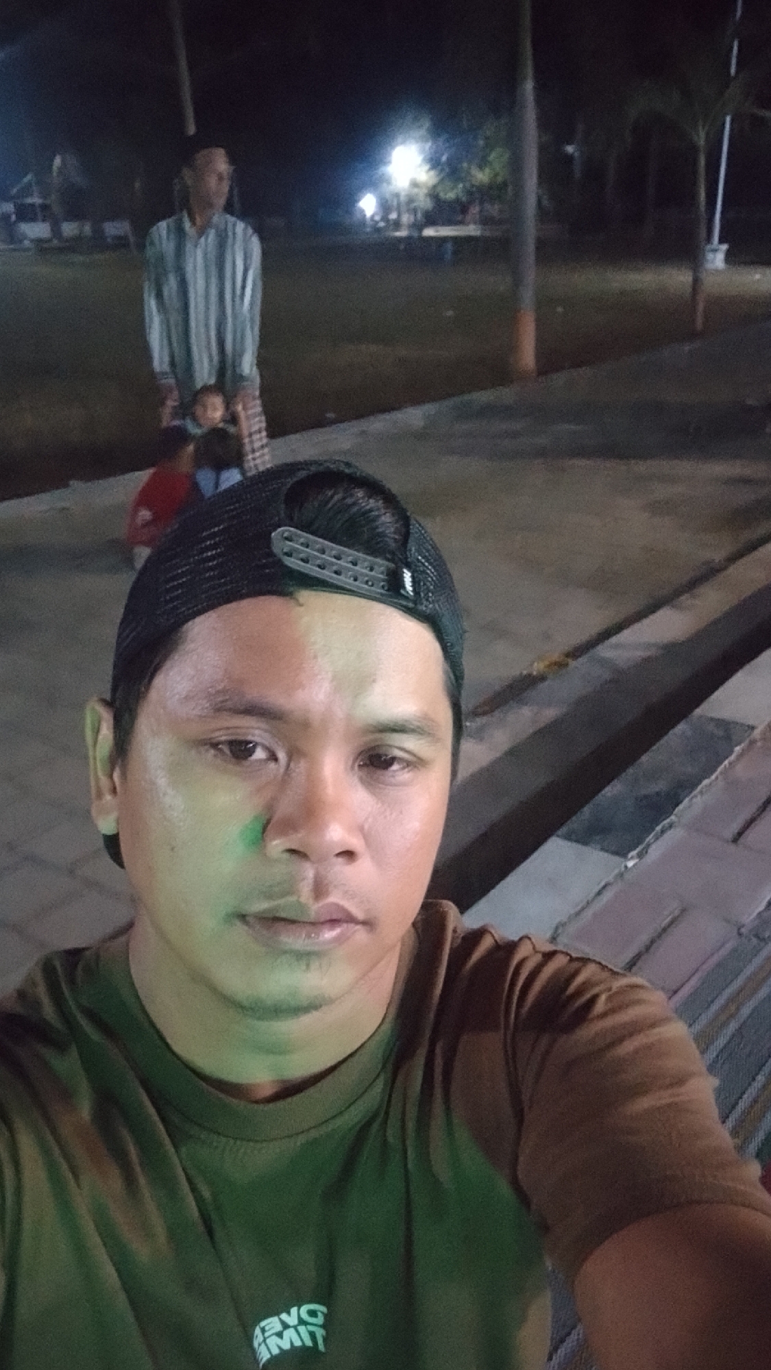 The image shows a selfie taken at night. The foreground is dominated by the face of a man wearing a dark baseball cap and a brown t-shirt. The t-shirt has white lettering on it that appears to be "MIT" and "ONE". The background is out of focus but shows an outdoor scene with a man and two children. The man in the background is wearing a striped shirt and a sarong or similar garment. One child is visible between the legs of the man, and another child is to the left of the man. There is a lamppost and some trees in the background. The scene appears to be a park or recreational area. The lighting suggests it is dusk or nighttime. Based on the information provided, the location is Karanglangu, Indonesia.