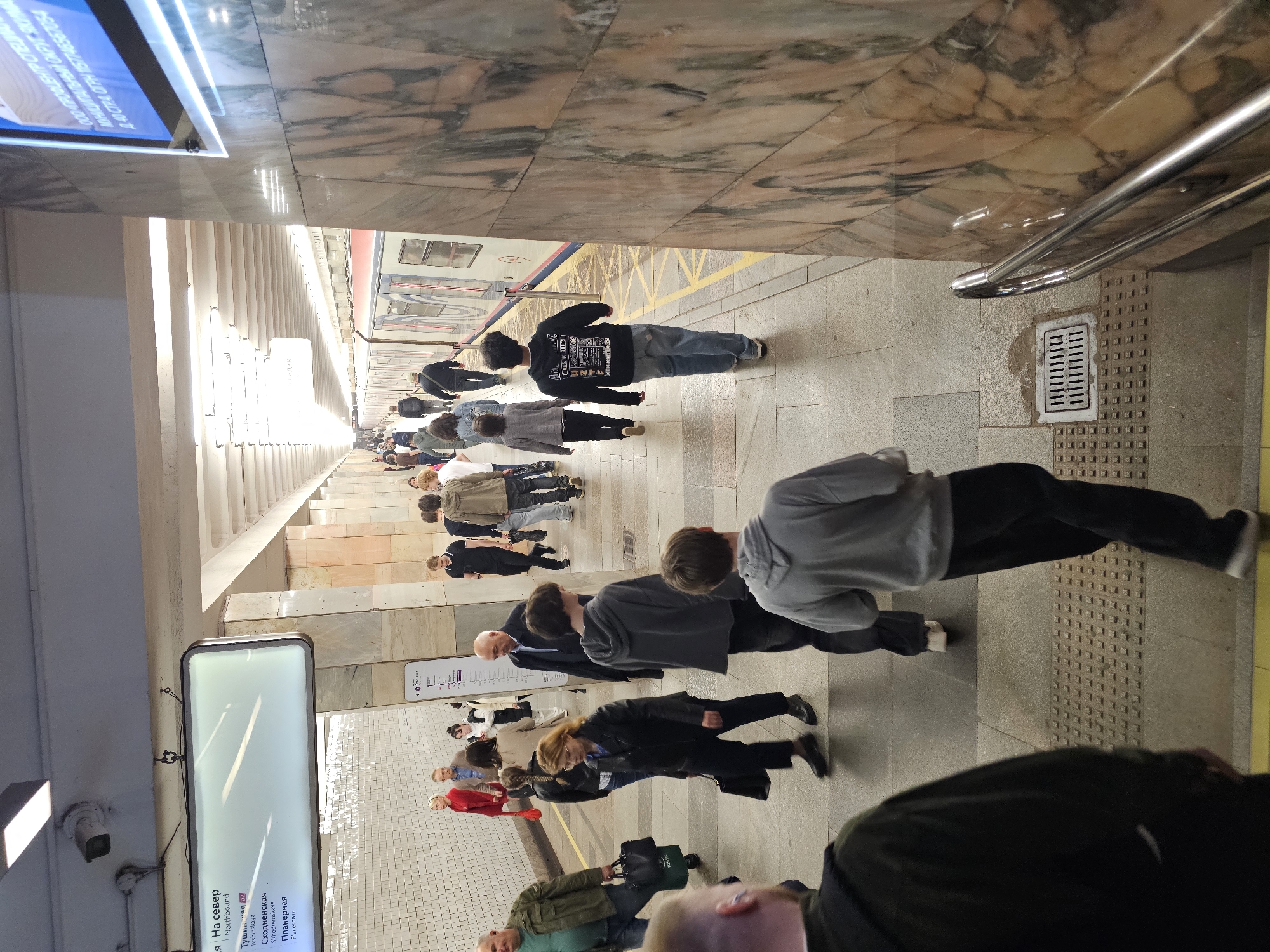 The image depicts a bustling subway station in Moscow, Russia, likely during peak hours. A queue of people waiting for the train is visible on the platform. There are commuters of different ages, styles, and attires. The architecture of the station showcases marble-clad walls and floors, typical of Moscow's metro stations. A digital information board indicates "Tushinskaya," "Skhodnenskaya," and "Planernaya" are the stations, signifying the train is heading north. The station is well-lit. A parked train is visible on the track.