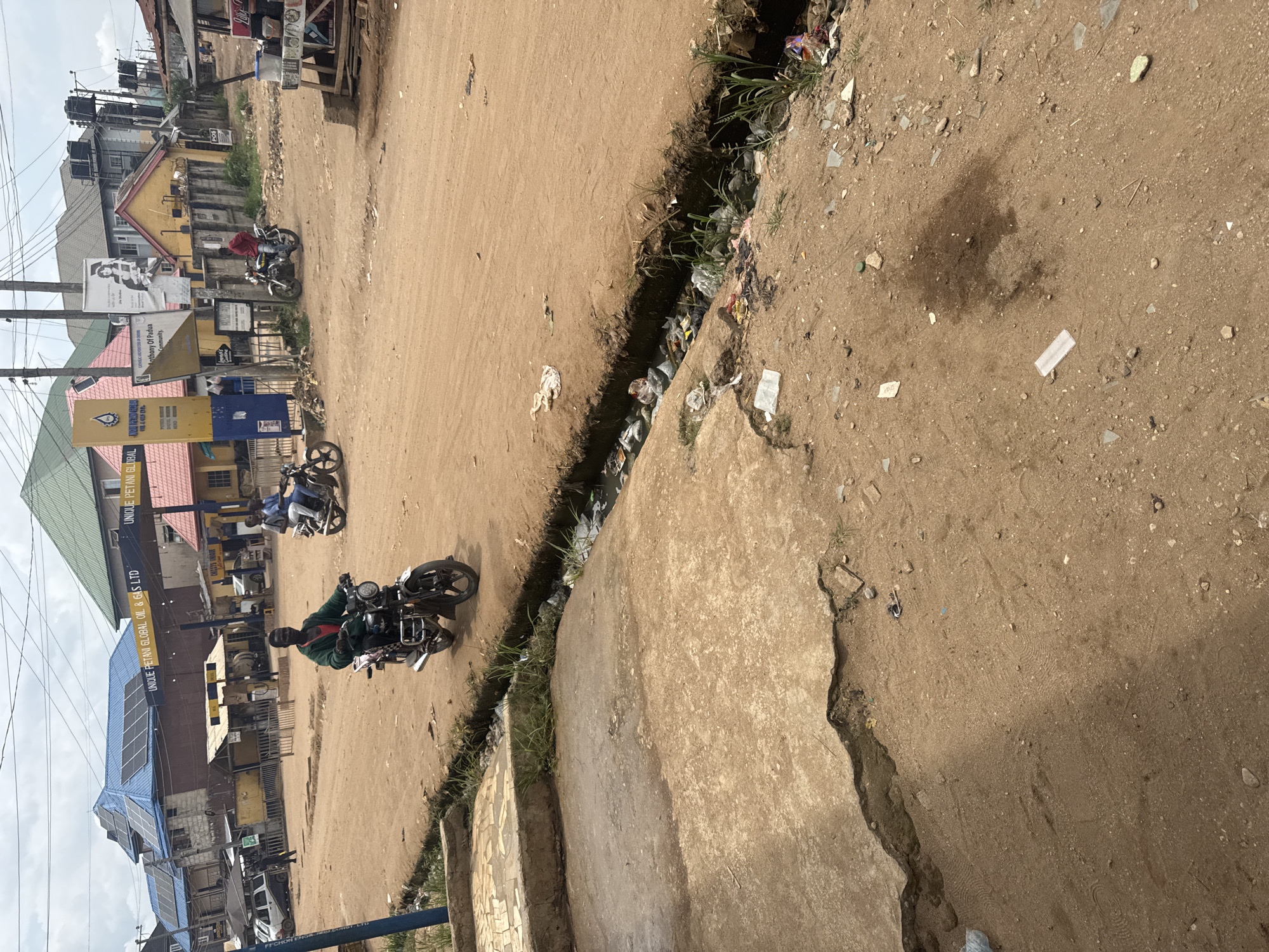 The image depicts a street scene in Ihiagwa, Nigeria. The road is unpaved, with a visible drainage ditch filled with trash running alongside a concrete sidewalk. Several motorcycles are present, one with a person riding it. Buildings line the street, including a gas station with signage that reads "UNIQUE PETANI GLOBAL OIL & GAS LTD". There are also signs that read, "Anthony Of Padua Community." Overhead power lines are visible. The area is dusty and shows signs of disrepair.