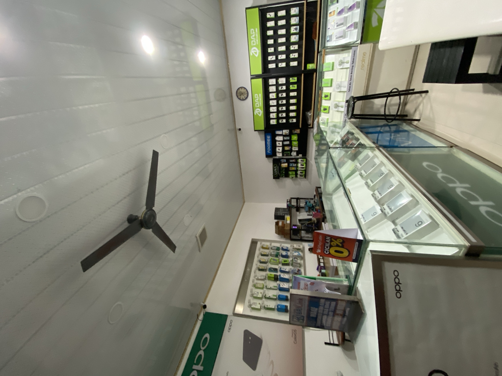The image depicts the interior of a brightly lit mobile phone store, likely located in Ta', Indonesia, given the brand "OPPO" prominently displayed and common in Indonesian markets. The store features a ceiling fan, fluorescent lighting, and white paneled walls and ceiling.
In the foreground, a glass display counter showcases various mobile phone accessories and devices. Signs for "OPPO" and "SPayLater CICILAN 0%" are visible, indicating promotional offers. Behind the counter, more display shelves are stocked with phone cases and accessories, with brands like "LUNA," "FOOMEE," "DAP," and "realme" clearly labeled.
To the left, a large advertisement for "OPPO" features an image of a smartphone. Further back, shelves are organized with a variety of phone accessories. A clock is mounted on the wall above the display shelves.
The overall environment suggests a retail space focused on mobile phone sales and accessories, with an emphasis on promotional offers. There are no individuals visible in the image.