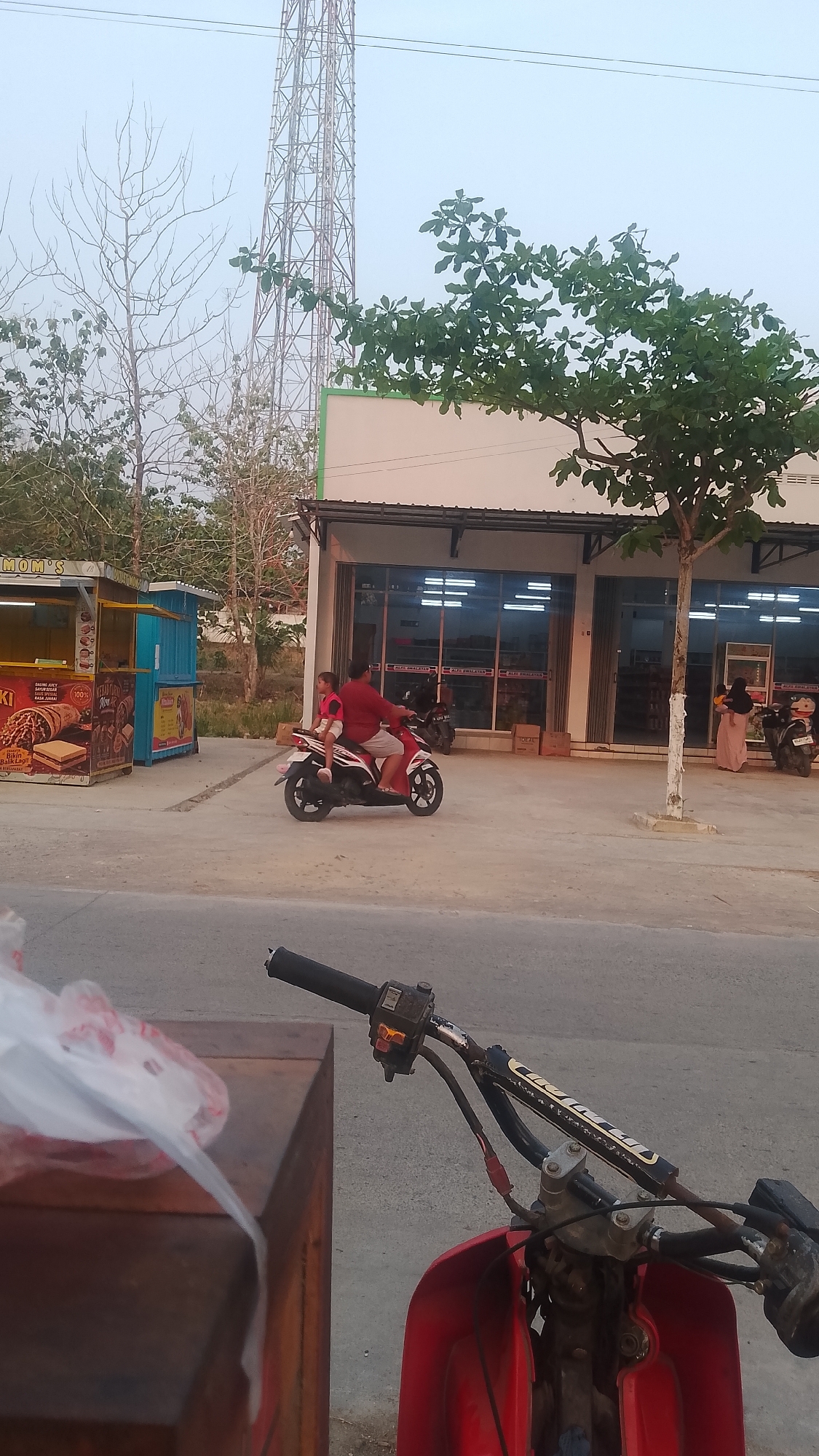 The image depicts a street scene in Karanglangu, Indonesia. In the foreground, the handlebars and front of a red motorcycle are visible, with a wooden structure to the left. In the midground, a man and a child are riding a red motorcycle. To the left of the motorcycle, there is a food stall with colorful signage. Behind the stall, a large telecommunications tower stands tall. In the background, a row of shops with large windows and awnings lines the street, with a woman holding a baby standing outside one of the shops. Trees and sparse vegetation are scattered throughout the scene. The sky is overcast.