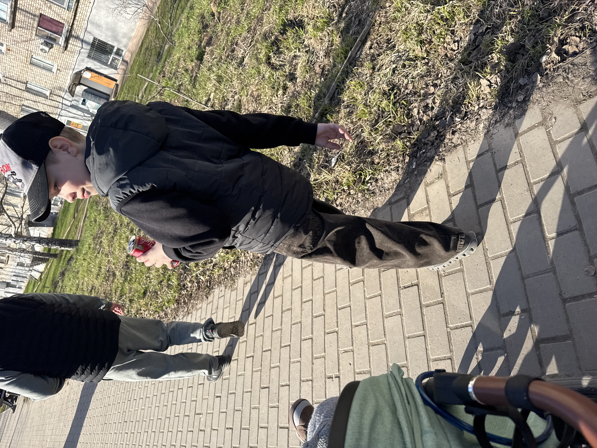 This outdoor image, likely taken in Chaikovsky, Russia, captures a sunny day scene on a paved sidewalk alongside a grassy area. The strong shadows indicate bright sunlight, possibly in the morning or afternoon. In the background, a multi-story brick residential building with numerous windows, air conditioning units, and security bars is visible, suggesting an urban or suburban setting. The trees are bare, pointing to early spring.
The primary subject is a young boy, fair-skinned with light hair, wearing a black quilted jacket, dark pants, and a grey and black baseball cap. He is walking on the paved path, looking slightly downwards, and holding a red can of Coca-Cola in his left hand. His expression appears neutral.
To the lower left, the legs and lower torso of another person are visible, wearing light gray sweatpants, dark sneakers, and a black vest over a long-sleeved shirt. Their posture suggests they are standing or walking near the boy. In the bottom right corner, handles resembling those of a stroller or pram, along with a piece of green fabric, are visible, indicating the perspective of the photographer or another individual pushing a child in a stroller. The feet of this third person, wearing brown shoes, are also partially seen in some crops.
The activity depicted is a casual stroll or outing, with the individuals moving along the paved path next to a patch of undeveloped ground. The visible text "Coca-Cola" is clearly identifiable on the red can held by the boy. The overall mood is relaxed and ordinary.