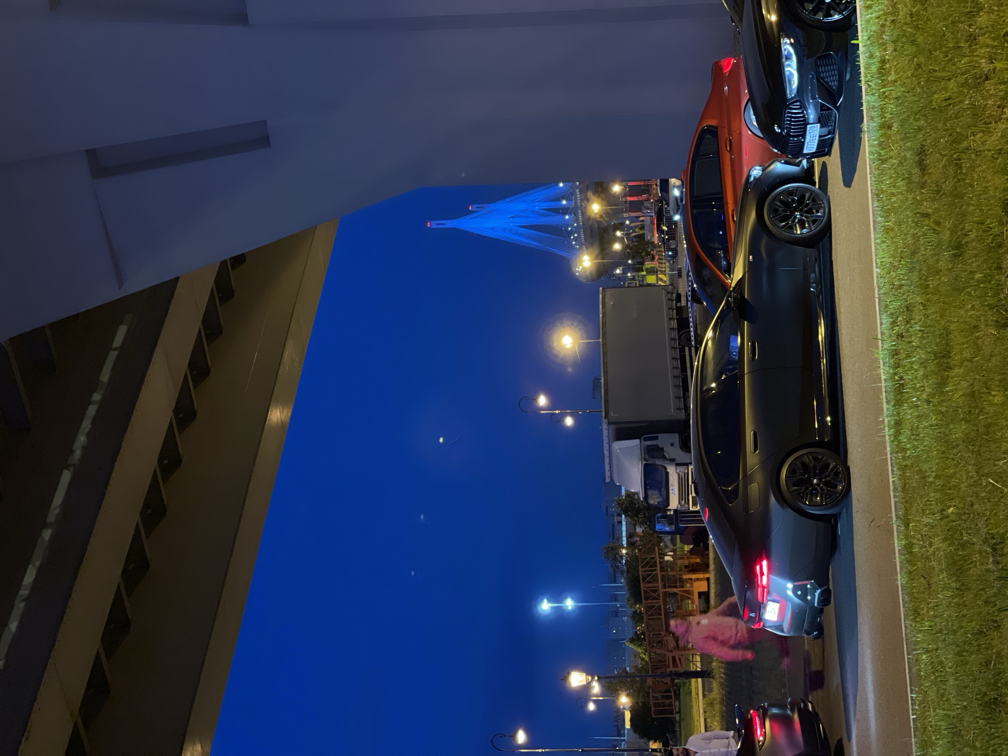 At dusk, a dark-colored BMW sedan with illuminated taillights is parked on the side of a road in an urban setting. The car is positioned on a paved surface, with a grassy median to its right. In the background, an orange sedan is also visible, along with other vehicles and a large truck. A prominent bridge structure, illuminated with blue lights, dominates the distant skyline. The scene is framed by the underside of an overpass in the foreground, casting a shadow over the area. Several streetlights are visible, casting a warm glow. Two men are present in the image; one is standing on the left, facing away from the camera, while the other is walking across the road in the mid-ground, his back to the viewer. The sky is a deep blue, indicating the transition from day to night.