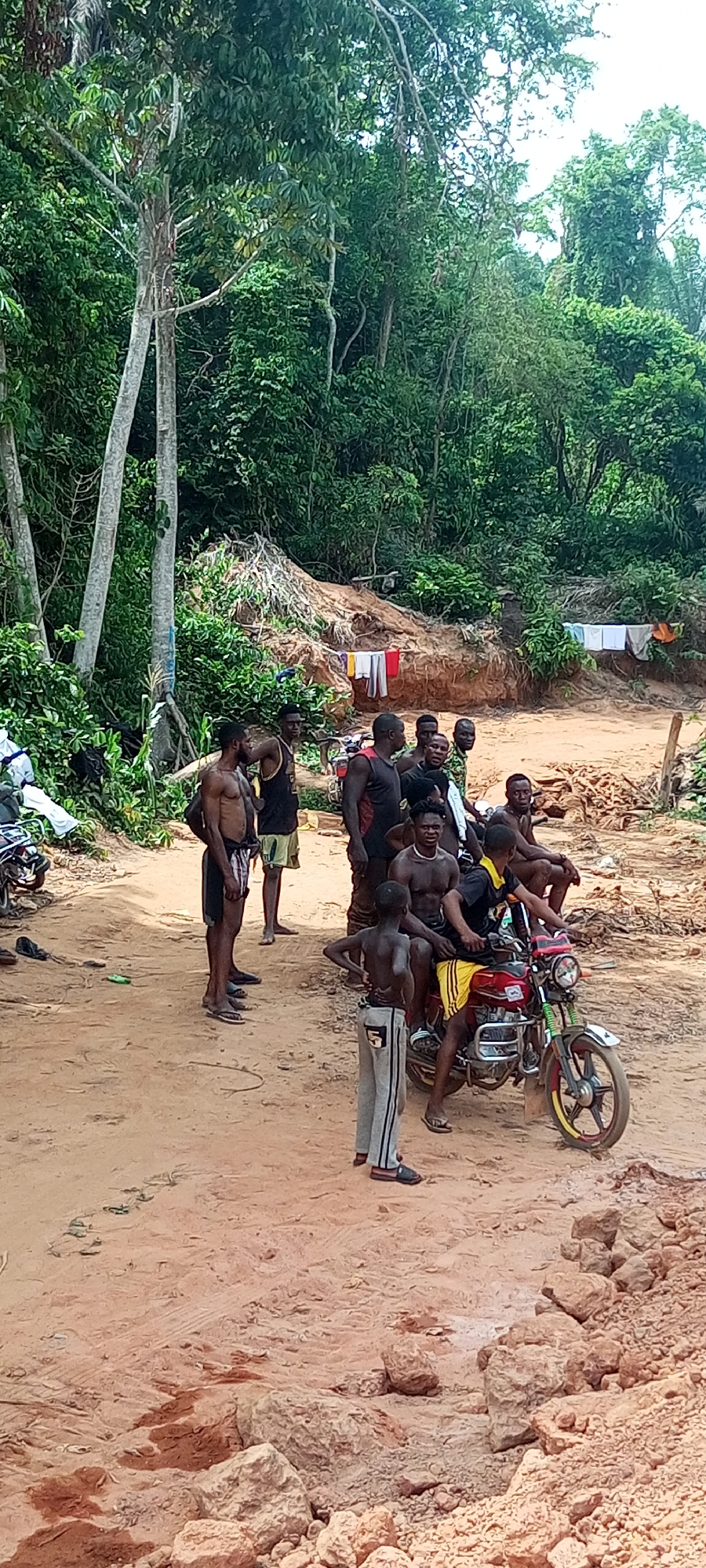 The image depicts a casual gathering of approximately 10-15 men and boys in a rural, forested setting in Aloko, Nigeria, during daylight hours. The ground is predominantly reddish-brown dirt, uneven and scattered with small rocks and debris.
In the midground, a man sits on a bright red motorcycle with distinct yellow accents on its wheels, wearing a black and yellow top and shorts. A young boy in grey sweatpants stands facing away from the camera, positioned in front of the motorcycle. Several other men, many shirtless or wearing tank tops and shorts, are standing and sitting around the area, engaging in what appears to be casual socialization or resting. Some individuals look towards the camera, with one man seated on the motorcycle displaying a relaxed smile.
The background features dense, vibrant green jungle vegetation and tall trees with light-colored trunks, indicating a heavily forested environment. A large mound of reddish-brown earth is visible in the mid-background, with some laundry hanging on a line or draped over it, suggesting a lived-in area. The lighting is bright and diffuse, consistent with an overcast or hazy day, and implies warm weather. No discernible text is visible within the image. The overall scene conveys a sense of a communal moment in a natural, unpaved outdoor environment.