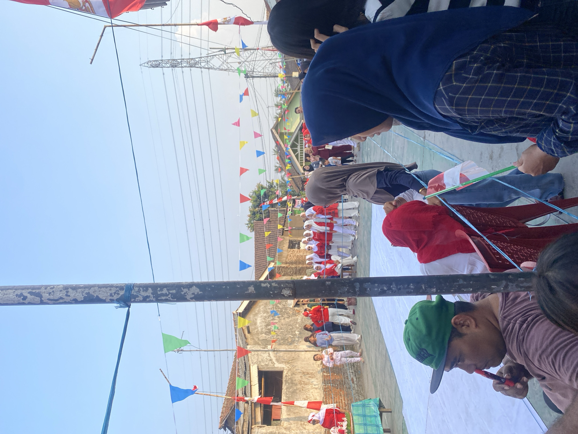 The image depicts an outdoor gathering, likely a community event, with numerous individuals present. The setting appears to be in a residential area of Cilegon, Indonesia, given the architecture of the buildings and the presence of a prominent electrical tower. A group of children and adults, many dressed in red and white attire, are gathered, suggesting a celebration, possibly related to Indonesian Independence Day. Decorative triangular flags in various colors are strung across the scene, enhancing the festive atmosphere. In the foreground, a man wearing a green cap is seated and holding a red and white object, possibly a small flag or a prop for a game. Several women wearing hijabs are also visible, some engaged in conversation or observing the event. The overall scene suggests a lively and communal celebration in an urban Indonesian setting.