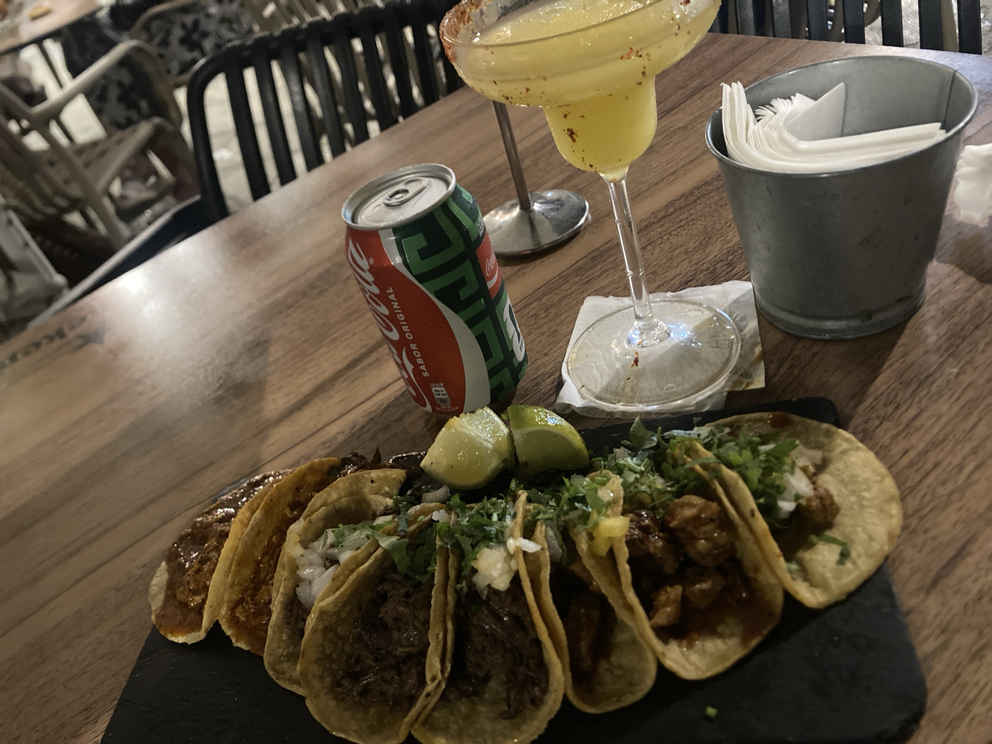 The image captures a detailed close-up of a dining table at a restaurant in Barcelona, Spain. On a dark, rectangular serving board, approximately seven tacos are arranged in a line. Several tacos feature corn tortillas filled with dark shredded meat, topped with finely diced white onions and chopped cilantro. Other tacos display a reddish, glossy sauce, indicative of birria. Two quartered lime wedges are positioned on the board among the tacos. Adjacent to the tacos, a red and green Coca-Cola can with "Sabor Original" printed in Spanish stands upright. To the right, a stemmed margarita glass, with a wide, chili-salted rim, contains a yellow-green beverage. This glass rests on a white paper napkin. Behind the margarita glass, the base of another stemmed glass is partially visible and out of focus. Further right, a small, galvanized metal bucket holds a stack of white paper napkins. The table surface is made of dark-stained wood planks, exhibiting subtle reflections. In the blurred background, several restaurant chairs are visible; some have dark slatted backs, while others are lighter in color with dark-patterned upholstery. No people are present in the scene.