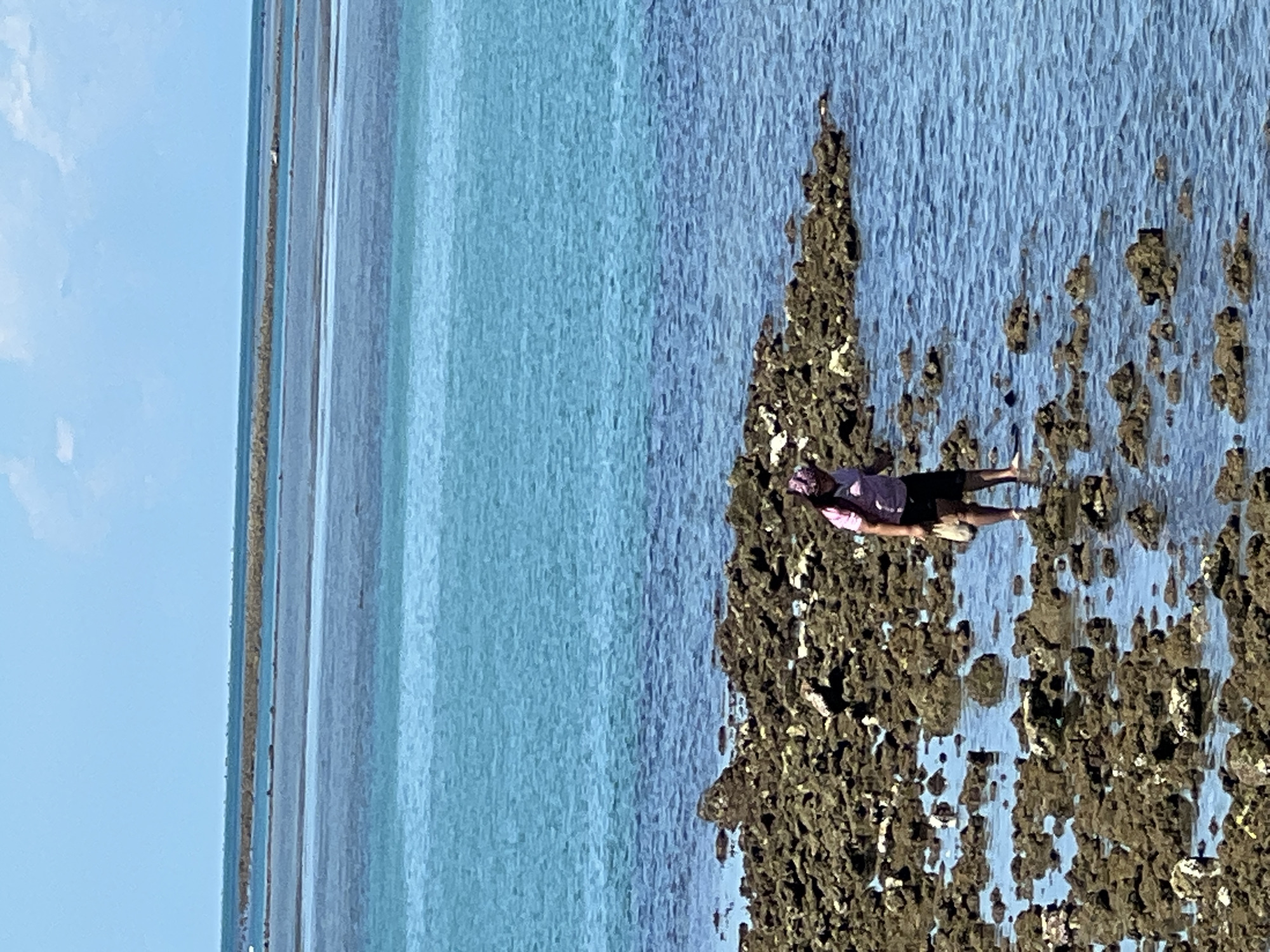 A person is walking through shallow water on a rocky shoreline, near Baan Hua Thanon, Thailand. The person, wearing a pink shirt and a patterned head covering, carries a light-colored bag. The water is clear and blue, revealing submerged rocks and the sandy bottom. In the background, there are distinct bands of water in varying shades of blue, suggesting shallow areas and a distant horizon. The sky is clear with a few scattered white clouds. The overall scene depicts a tranquil coastal environment.
