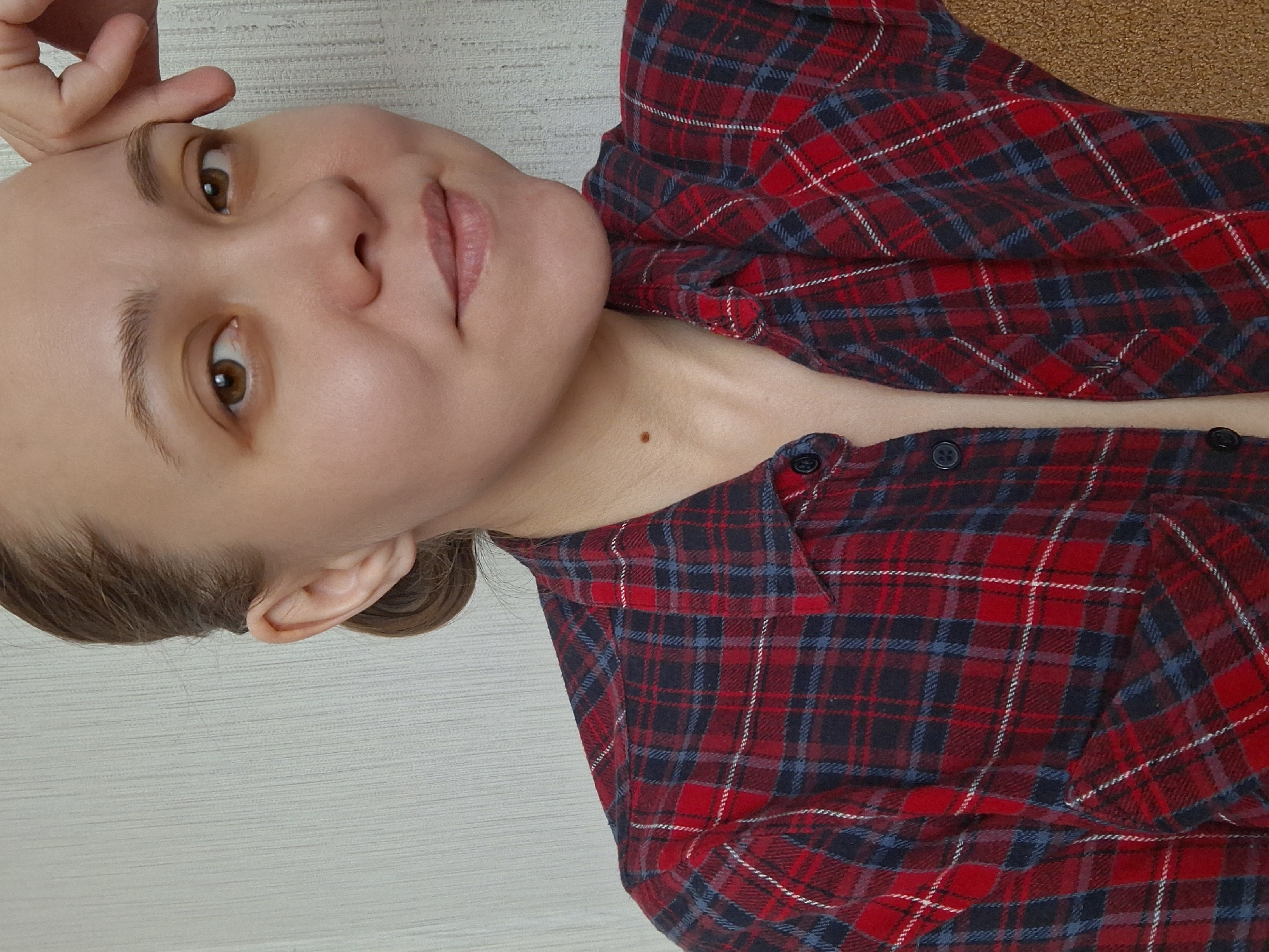 A woman is facing the camera, wearing a red and blue plaid shirt with a few top buttons undone. She is indoors, possibly at a home or apartment. The background is a light, textured wall, and there's a hint of a corkboard-like surface behind the shirt. Her hair is pulled back. It is not possible to verify the location of Chaikovsky, Russia.