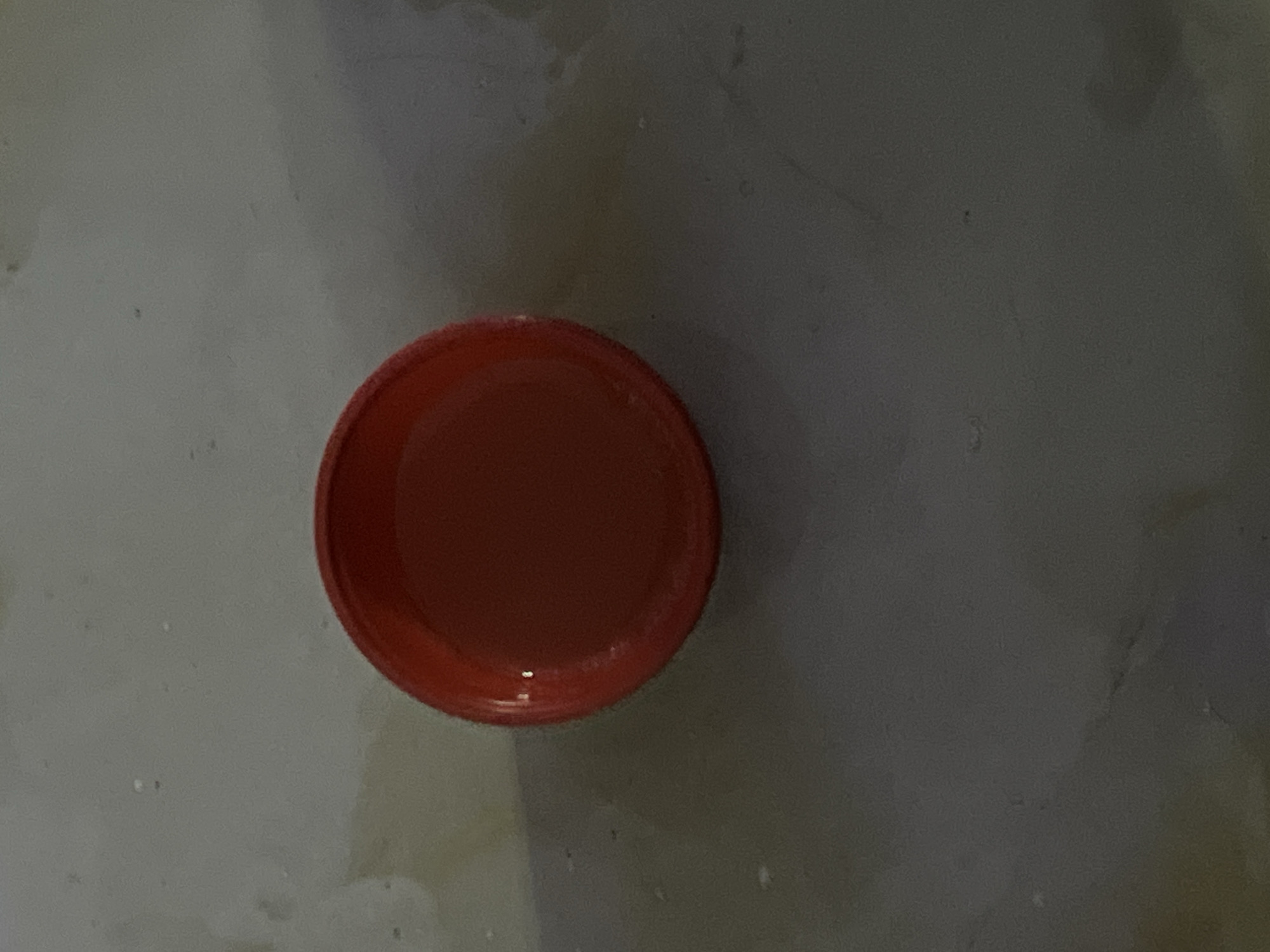 A red bottle cap is centered on a light gray, textured surface. The cap is round with a slightly raised rim and appears to be made of plastic. The surface beneath the cap is uneven and shows some discoloration, with lighter patches and subtle shadows. The lighting is dim, casting a soft glow on the cap and creating a muted atmosphere. There is no indication of the location in Jauro Yinu, Nigeria within the image itself.