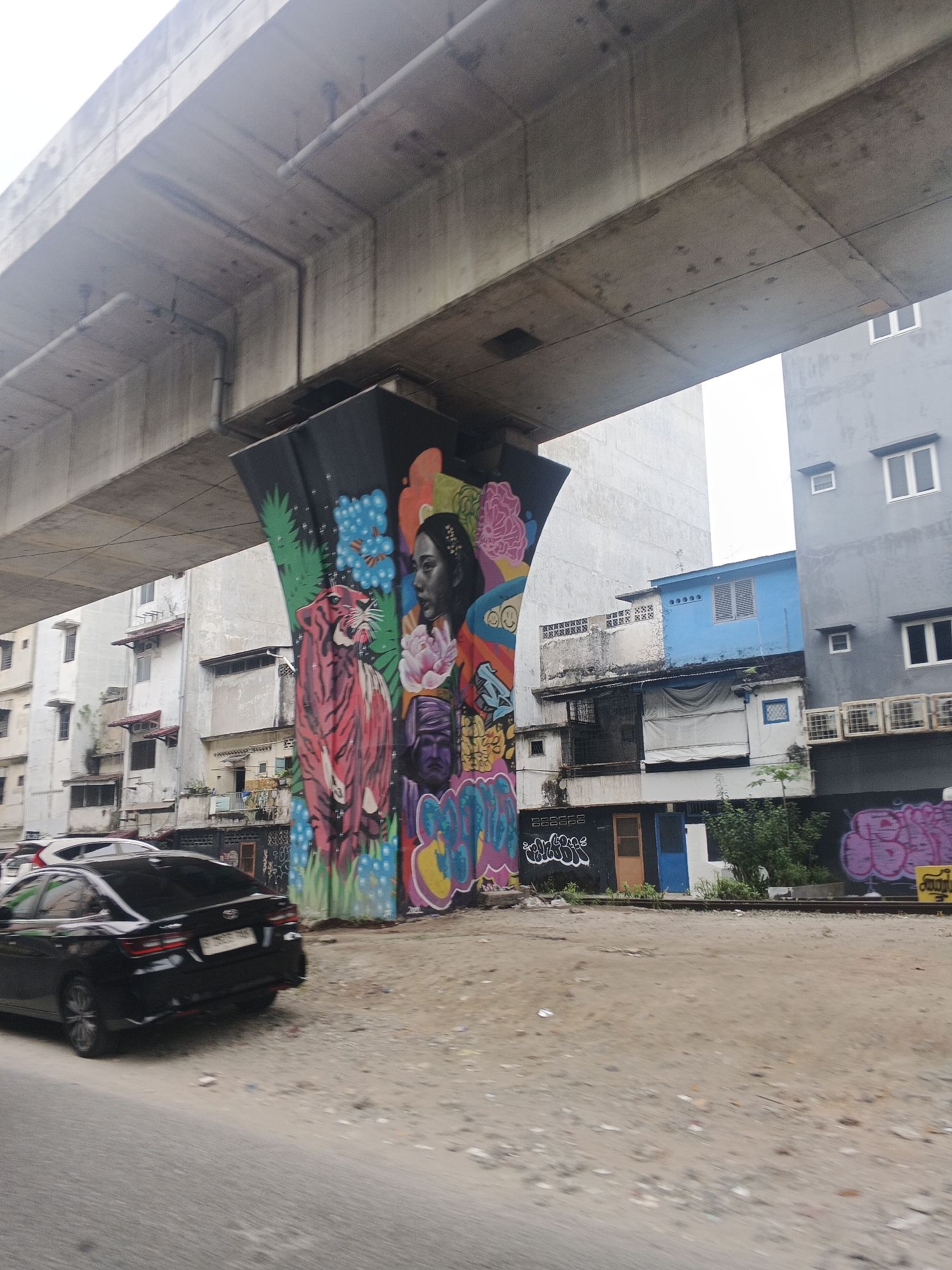 An outdoor scene captures a street-level view beneath an elevated concrete structure, likely a highway or overpass, in the City of Medan, Indonesia. A large concrete pillar supporting the overpass is adorned with vibrant graffiti art. The artwork features a stylized pink tiger with green foliage, a portrait of a woman with dark hair, and various abstract shapes and lettering in bright colors.
Behind the graffiti-covered pillar, a row of buildings is visible. The closest building to the left is a light-colored, multi-story structure with windows. To its right, a narrower, older building exhibits signs of age, with some covered windows and balconies. Further to the right, a bright blue, two-story building stands out, with windows and what appear to be air conditioning units. Next to the blue building is a gray, multi-story building with several rectangular windows and external air conditioning units.
In the foreground, a black sedan is partially visible on a road, indicating a street or pathway. The ground in front of the buildings and beneath the overpass is a dirt and gravel area with some debris. Railway tracks run horizontally across the mid-ground, suggesting the presence of a train line. The sky is overcast and appears bright.