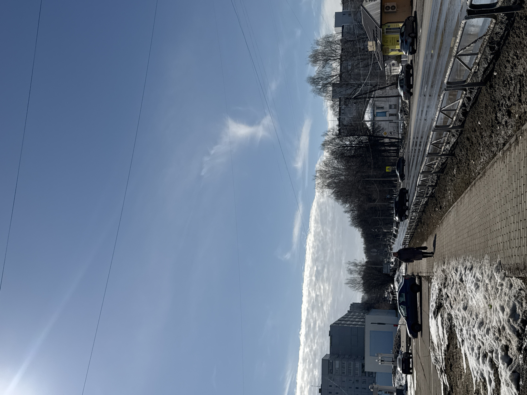 This image depicts an urban street scene in Chaikovsky, Russia, observed during daytime under a bright blue sky with scattered clouds and visible utility wires. The sun, partially visible in the upper left, casts a lens flare.
In the foreground, a brick-paved sidewalk extends from the bottom center towards the middle distance. To the left of the sidewalk, patches of melting snow and exposed earth are present. A single individual, dressed in a dark coat, walks away from the camera on the sidewalk, carrying a dark bag in their left hand. To the right of the sidewalk, a metal barrier separates it from a wet asphalt road. Mud and slush are visible on the ground between the barrier and the road.
The midground features a multi-lane road with several vehicles, both moving and parked. The road surface appears wet, likely from melted snow. On the left side of the road, multiple gray multi-story residential buildings line the street. On the right side of the road, behind the barrier, are more bare deciduous trees and various structures, including a distinctive yellow one-story building with a dark roof and external air conditioning units. Further down the street on the right, another white building with a blue facade is visible.
In the background, more urban structures and bare trees are visible under the partially cloudy sky.