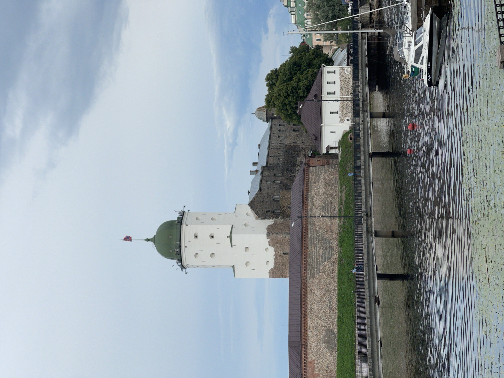 The image depicts Vyborg Castle, a medieval fortress located in Vyborg, Russia, situated on the Vyborg Bay. The central focus is the prominent white tower of the castle, topped with a green dome and a flag. Adjacent to the white tower is a darker, stone-clad section of the castle complex. In the foreground, a stone embankment is visible, leading to a bridge that spans a body of water. The water is calm, with visible ripples and a scattering of water lilies. Several boats are moored near the bridge, including a white motorboat with "Eagle" visible on its side and a sailboat. A few individuals can be seen walking on the bridge. The sky above is overcast with dramatic, dark clouds. The scene conveys a historical and somewhat somber atmosphere, characteristic of an ancient fortress.