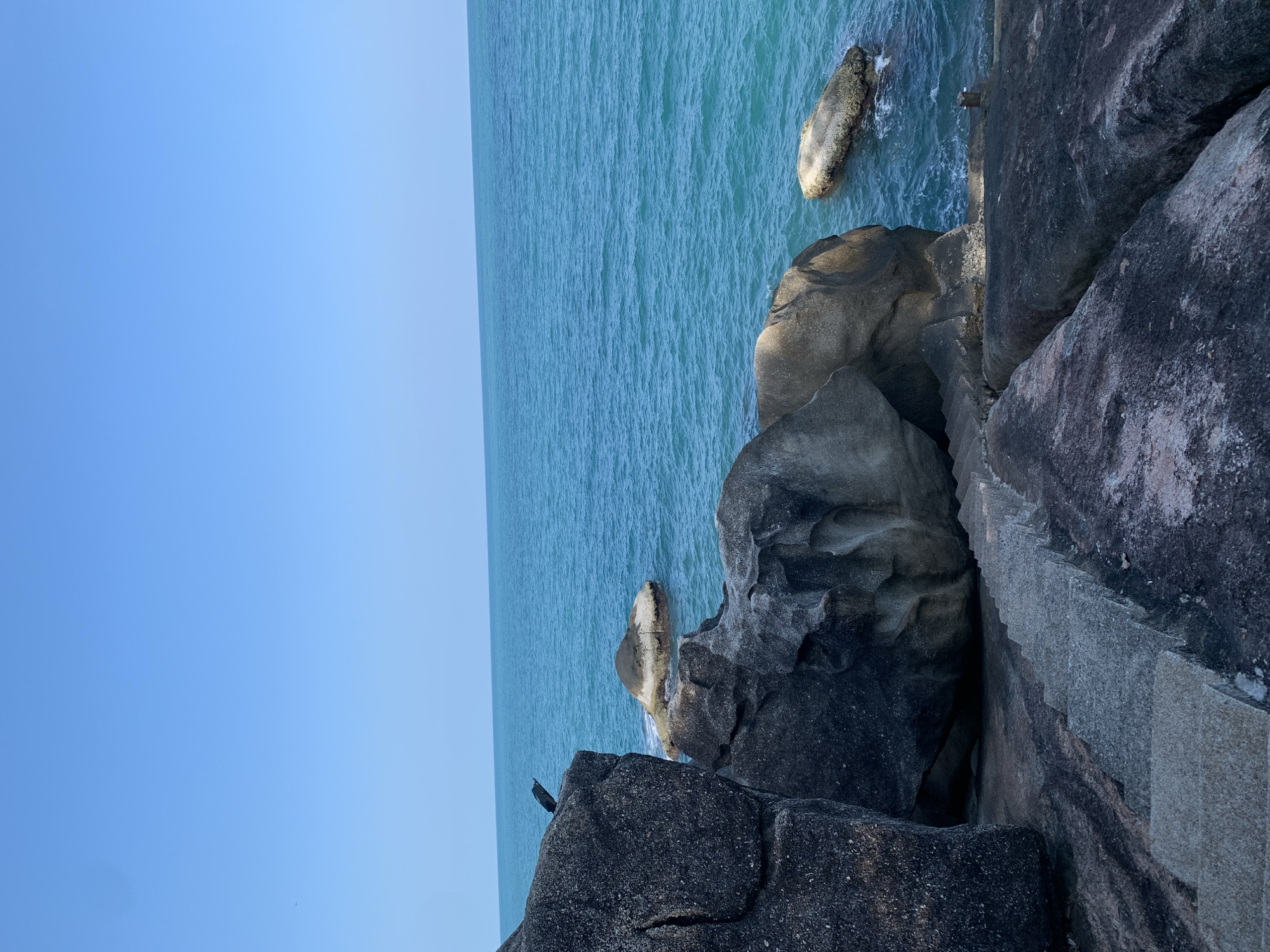View of the coastline in Baan Chaweng Noi, Thailand, featuring rock formations and stairs leading down to the water. The water is turquoise and the sky is light blue. A small, dark-colored bird is perched on the rocks.