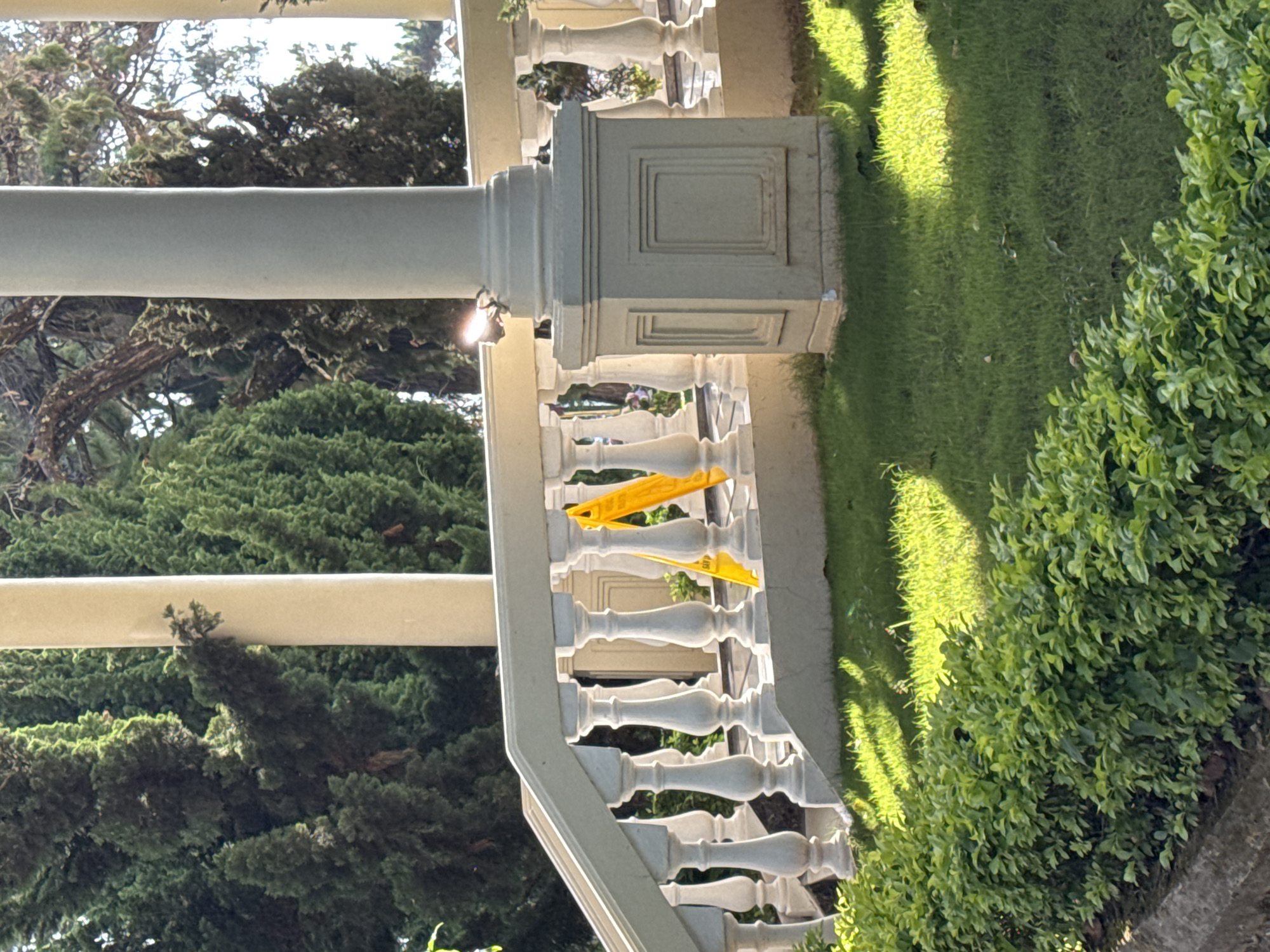 An eye-level, horizontally-oriented shot presents a segment of a building exterior, specifically the balustrade of a balcony, potentially in Da Lat, Vietnam. The building is likely made of concrete or stone, given its texture and color.
The balustrade, painted white, features classical detailing, with individual balusters arranged in a repeating pattern along the balcony. A yellow object, perhaps a piece of plastic or fabric, is draped over the railing, adding a splash of color.
Behind the balustrade is a verdant landscape, composed of varied shrubbery and trees. The sunlight filters through the foliage, casting shadows on the balcony and the surrounding vegetation. The ground is covered with grass.