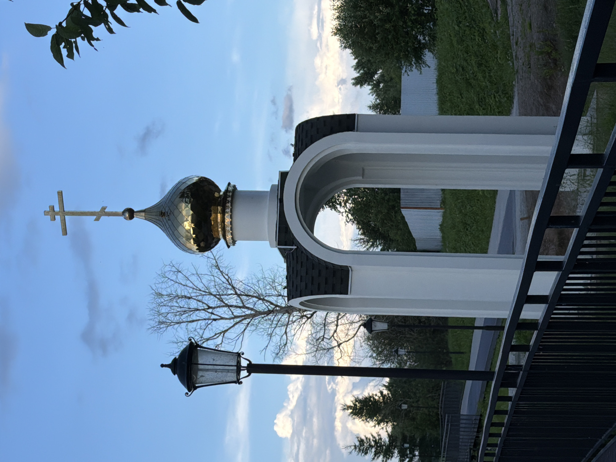 This image, taken in Solikamsk, Russia, captures a serene outdoor scene dominated by the architectural features of a religious building and its surroundings. The primary subject is a domed structure, likely a church or chapel, distinguished by its golden dome topped with a cross. The dome appears ornate, reflecting the ambient light. Adjacent to it is a white architectural element with a rounded arch, possibly part of an entryway or pavilion.
In the foreground, a traditional lamppost with a glass lantern stands on a dark pole, adding a touch of historical charm. The background reveals a lush green lawn, dense trees, and a fence, suggesting a well-maintained park or religious grounds. The sky is a clear blue, dotted with scattered clouds, indicating fair weather. A bare-branched tree is visible behind the lamppost, its silhouette contrasting with the foliage.
The time of day appears to be either early morning or late afternoon, given the soft lighting and the gentle illumination of the clouds. No people are visible in the frame, creating a sense of peaceful solitude. The overall mood is tranquil and contemplative. There is no visible text in the image.