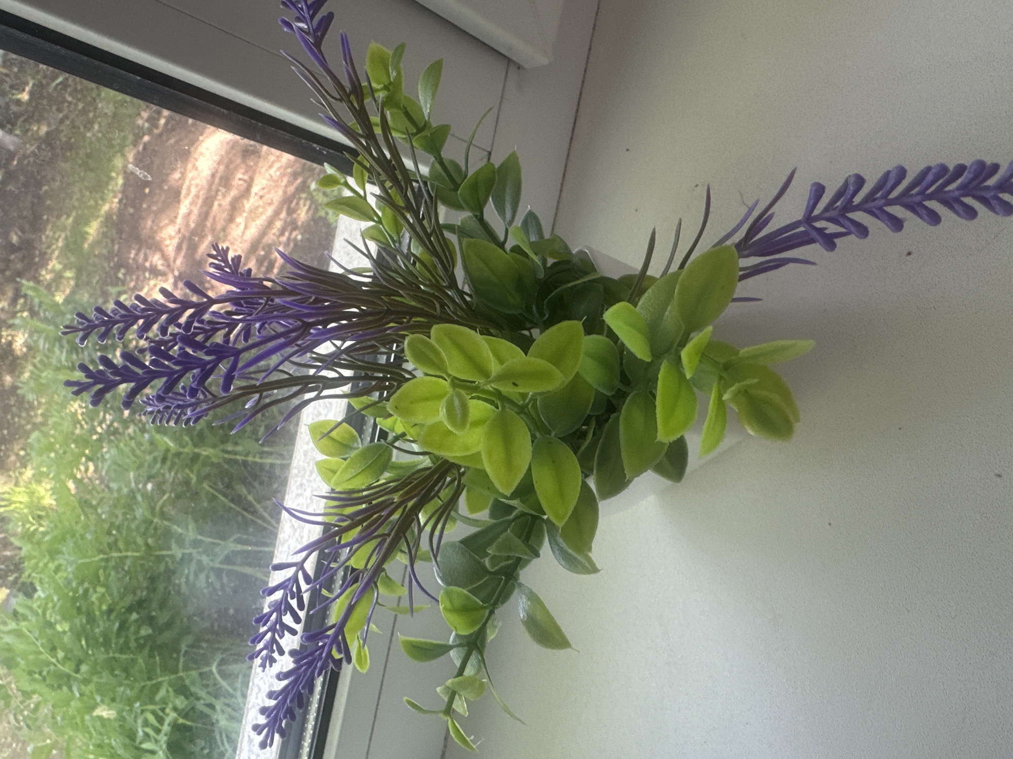 A close-up shot shows a faux bouquet of lavender and green foliage resting on a white windowsill. The artificial lavender stems are a vibrant purple with delicate, textured florets, interspersed with thin, dark green leaves. The green foliage consists of clusters of small, oval-shaped leaves in varying shades of light and medium green, with some stems drooping downwards.
Through the window on the left, a blurry background suggests an outdoor scene with lush green trees and vegetation, hinting at a daytime setting. The light filtering through the window casts a soft glow on the windowsill and the artificial plants, highlighting their texture and form. The overall impression is one of simple, decorative arrangement on a windowsill, possibly in a home or office. No people are visible in the image. No text is present.