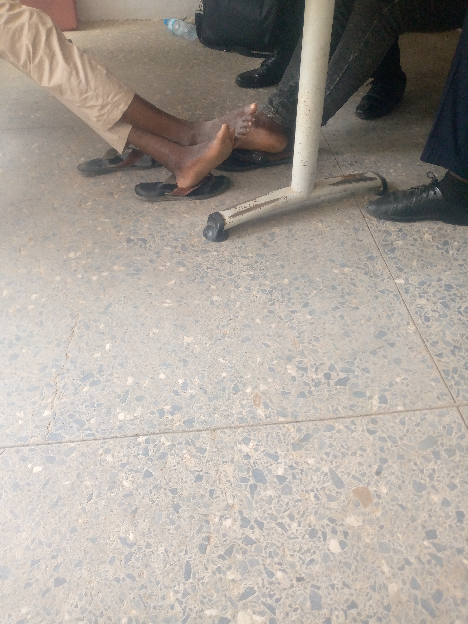 The image shows the lower bodies of at least three people sitting or standing around a white metal table leg on a terrazzo floor. One person's legs are extended, wearing light-colored pants and black flip-flops. Another person's legs are visible behind the table leg, wearing dark pants and black dress shoes. A third person's legs and feet are visible on the right, wearing dark pants and black dress shoes. The floor is a speckled grey terrazzo with visible grout lines. In the background, a clear plastic water bottle and a dark bag are present. The scene appears to be indoors, possibly in a waiting area or common space, with artificial lighting. There are no obvious indications of the specific location within Jalingo, Nigeria, beyond the general setting.
