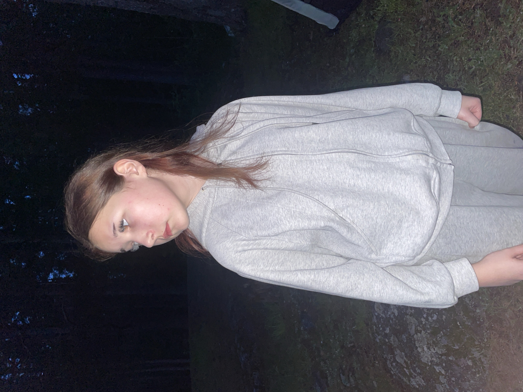 A young person, possibly a girl, is shown in a portrait-oriented, full-body shot. She has light skin, long reddish-brown hair, and is wearing a light grey hooded sweatshirt and matching sweatpants. Her face is visible in profile, looking downwards and slightly to her right, with a somewhat somber or pensive expression. Her left hand is clenched into a fist at her side, and her right hand is partially visible further down.
The setting appears to be outdoors, likely at night or in very low light conditions, given the dark background. There are hints of foliage or trees in the background, suggesting a natural environment like a forest or park. Patches of green grass are visible beneath her feet. The overall lighting is dim, with some faint light illuminating her face and the front of her clothing, possibly from an artificial source or residual twilight. There is no visible text in the image.