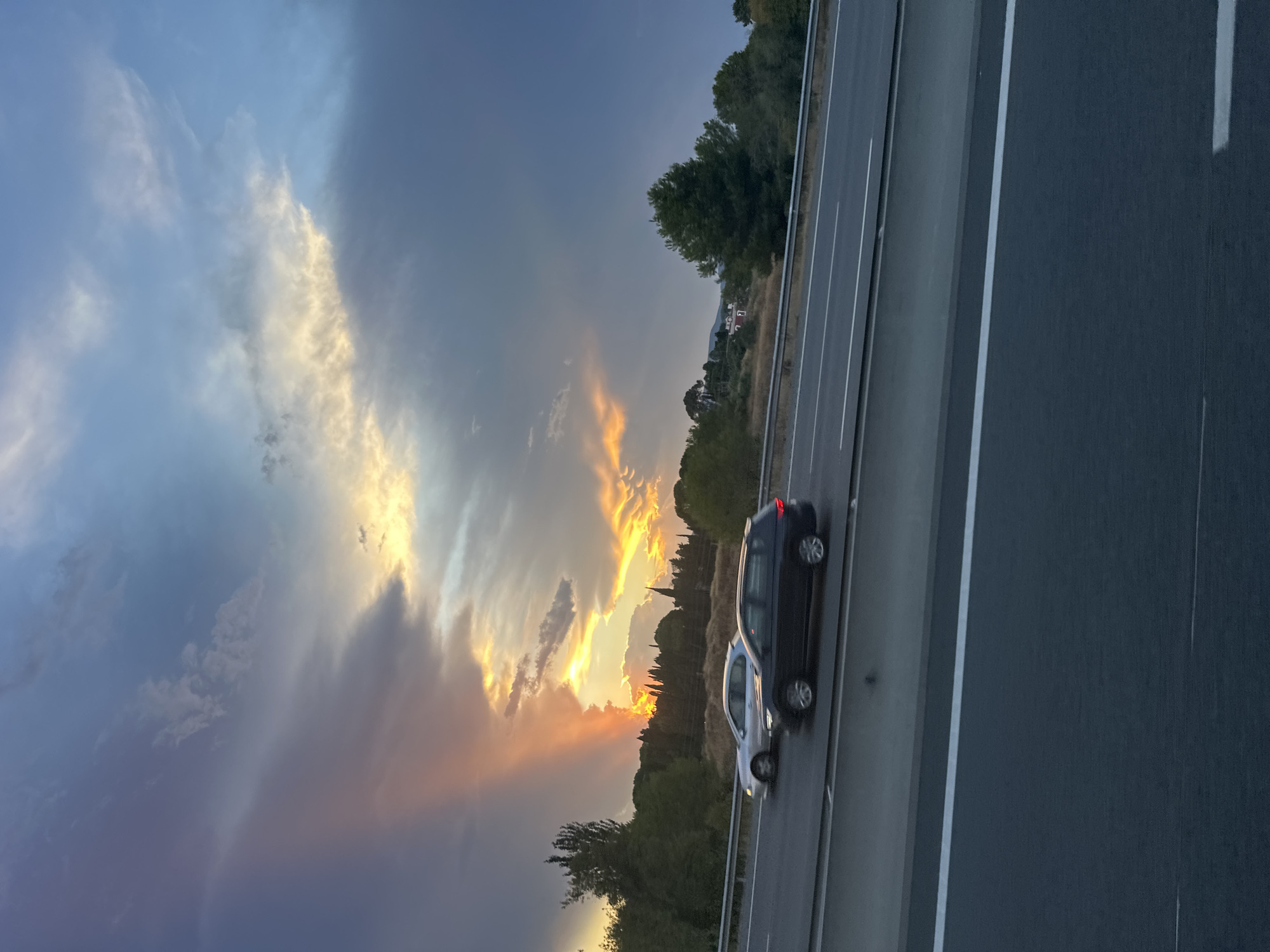 The image depicts a highway scene near La Granada, Spain, during what appears to be sunset or a similar atmospheric event. The sky is filled with dramatic clouds, with streaks of orange and yellow light breaking through, suggesting a colorful twilight. A narrow, bright shaft of light, possibly from a sunbeam or a localized weather phenomenon, pierces through the darker clouds on the left side of the frame, illuminating the sky in that area with a vibrant reddish-orange hue.
On the highway, two cars are visible. The car in the foreground is a dark-colored SUV, possibly a wagon, driving in the right lane. To its left, in the adjacent lane, is a silver sedan. Both vehicles are in motion.
The highway itself is paved and multi-laned, with white lines marking the lanes. A metal guardrail separates the highway from the embankment on the left.
Beyond the highway, a landscape of trees and vegetation can be seen. The trees are densely packed in some areas, with some taller, darker trees on the left and more scattered evergreen trees on the right. In the middle ground, a few houses with red roofs are nestled amongst the trees on a hillside, suggesting a residential area bordering the natural landscape. The overall impression is of a semi-rural or suburban setting on the outskirts of a town, with the dramatic sky adding a striking element to the everyday scene of a highway.