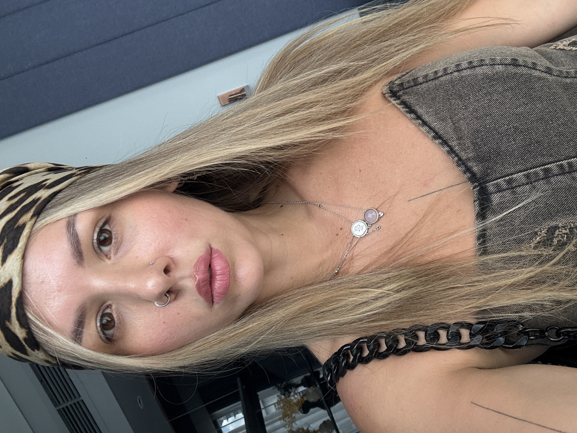 A woman is taking a selfie in what appears to be a room. She has long blonde hair and is wearing a leopard-print headband, a nose ring, and a denim top. She is also wearing a necklace with two pendants. The background is out of focus but appears to be a room with some shelving units visible.