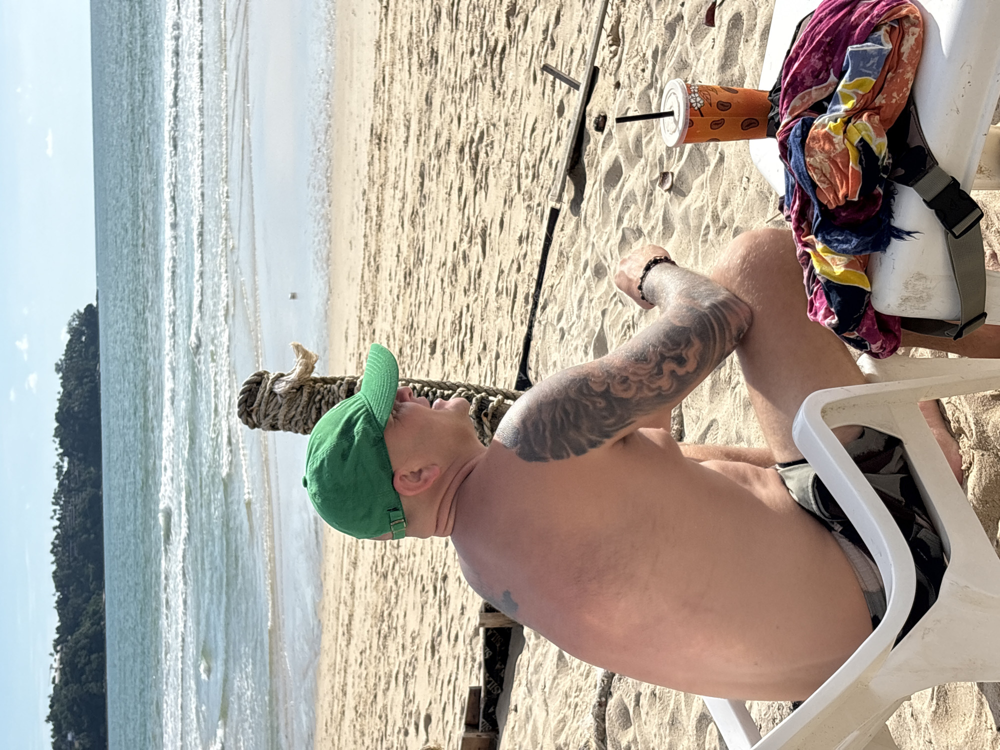 A man with tattoos on his back and left arm sits in a white plastic chair on a sandy beach, facing right. He wears a green baseball cap and camouflage-patterned swim trunks. In front of him, on a white table, is an orange beverage in a clear plastic cup with a black straw, and a pile of colorful folded fabrics. Behind the table, a thick rope is tied to a wooden post. The ocean with gentle waves and a clear sky are visible in the background. The beach is composed of light-colored sand. The man is on a beach in Choeng Thale, Thailand.