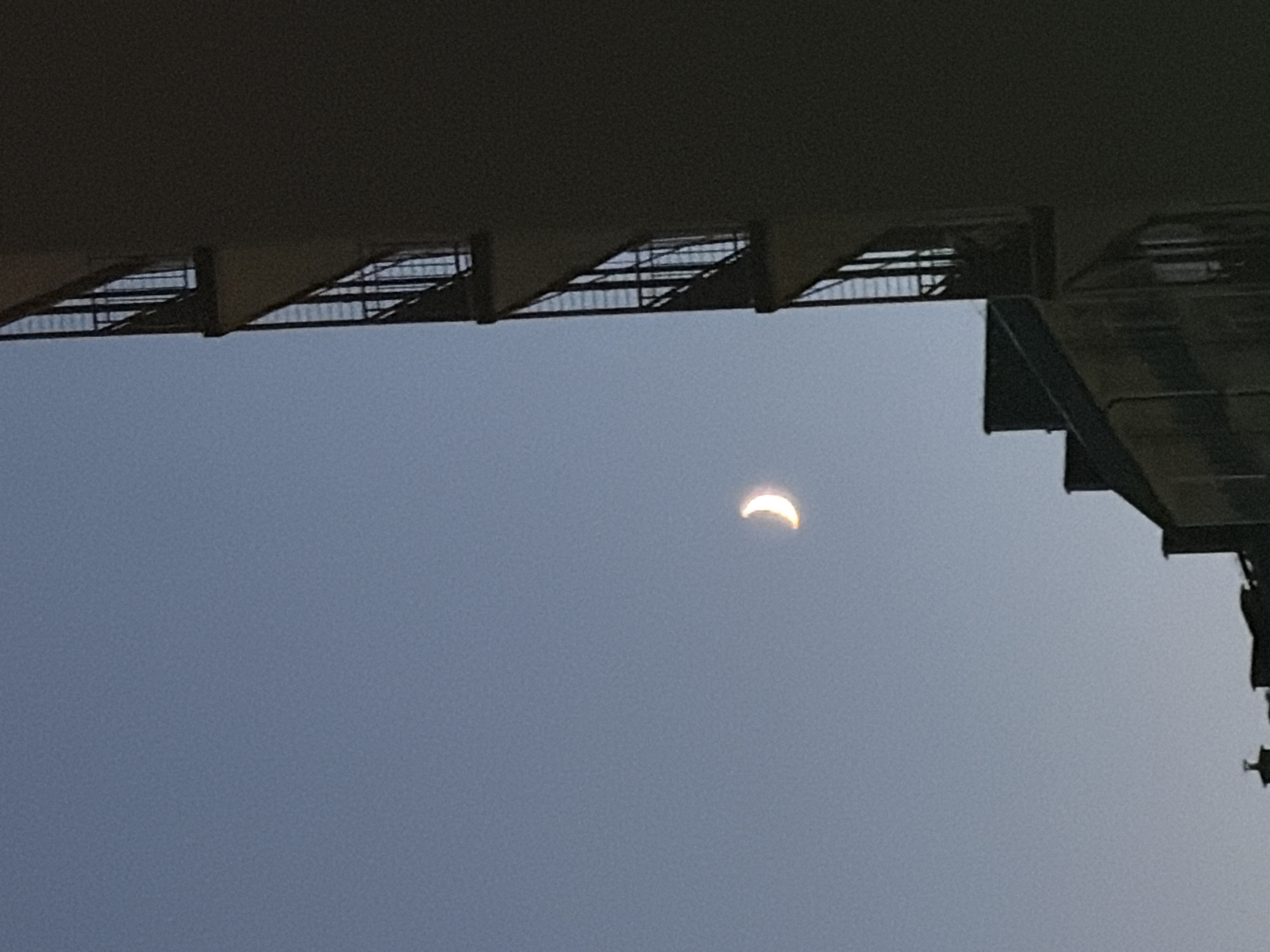 A crescent moon is visible in a clear, pale blue sky. To the right of the moon, a tall, dark building with balconies occupies the right side of the frame. The building is silhouetted against the sky, with the balconies appearing as dark rectangular openings with railings. In the lower portion of the image, the rooftops of other buildings are visible, also in silhouette. The scene appears to be from an urban environment, captured during twilight or early evening.