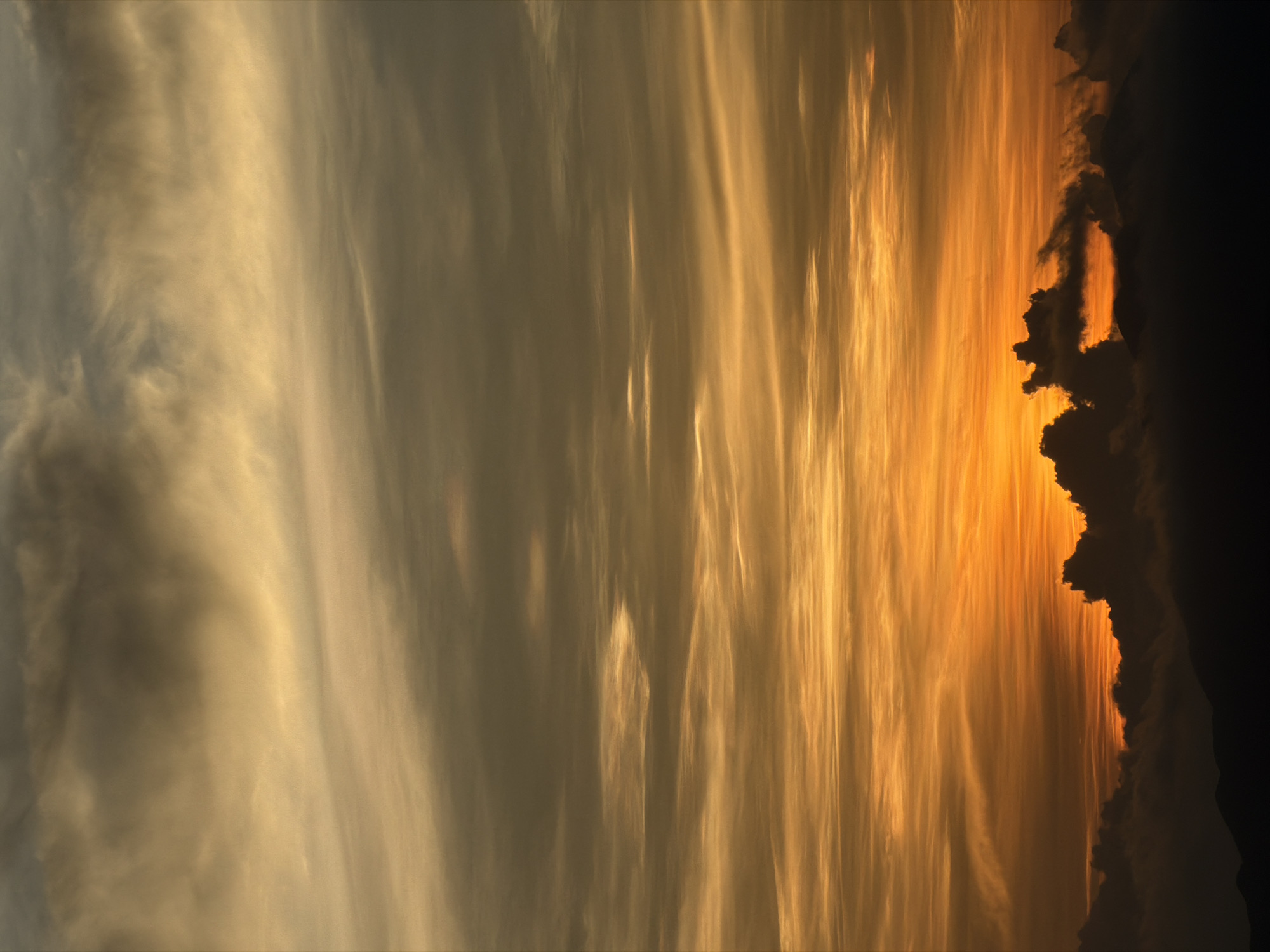 A vertical, medium shot captures a dramatic sunset over a mountainous landscape. The sky is filled with wispy, striated clouds, transitioning from a bright, fiery orange near the horizon to a muted yellow and then to a darker, grayish-brown at the top. The lower portion of the image is dominated by the silhouettes of dark, undulating mountains and thick, billowy clouds, obscuring the terrain below. The sunlight pierces through a gap in the clouds at the horizon, creating a vibrant orange glow. The scene is devoid of people or any identifiable man-made structures, suggesting a remote, natural setting. The lack of specific landmarks or context makes it impossible to pinpoint a location within Hoi An, Vietnam. The image focuses solely on the atmospheric and geological elements of the landscape.