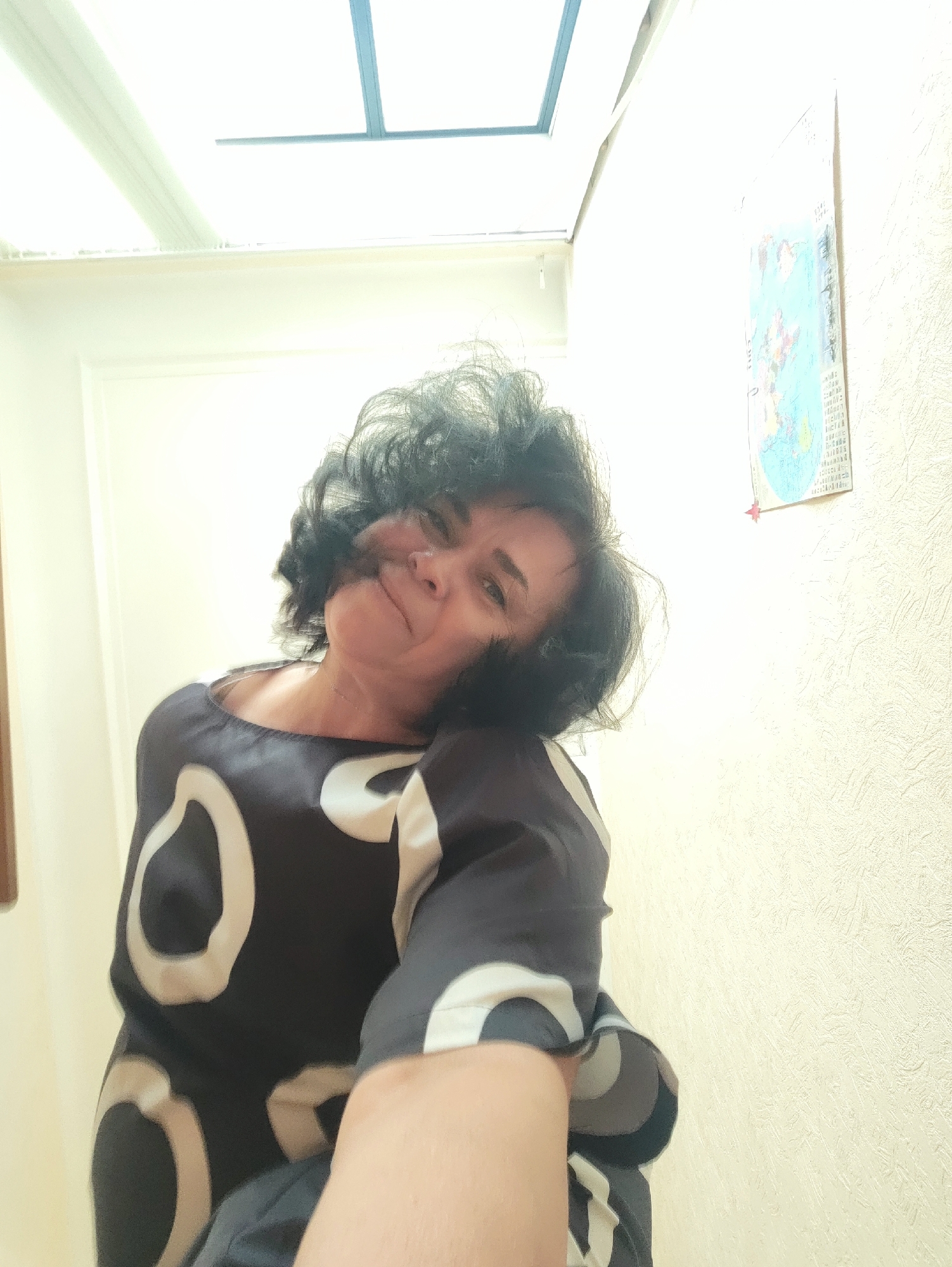 The media file shows a selfie of a woman with dark, curly hair. She is wearing a dark top with a pattern of white and light gray circles. The woman is leaning her head to the side and smiling slightly, with her eyes looking towards the camera. The lighting suggests it is daytime, with a skylight visible in the background. A textured wall is on the right side of the image, with a small, colorful chart or poster pinned to it. The overall setting appears to be indoors, possibly a home. There is no discernible text in the image.