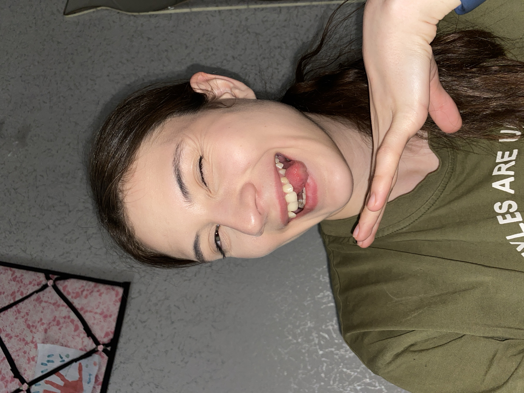 A young woman with long, dark brown hair is lying on her back, presumably on a gray floor, wearing an olive-green t-shirt. Her face is partially obscured by her own hand, which is positioned near her hair. She has a mischievous expression, with one eye closed and her tongue slightly protruding. To the left of her head, there is a framed object with a pink and white geometric pattern and a handprint visible on a piece of paper within the frame. No information suggests this image was taken in Votkinsk, Russia.