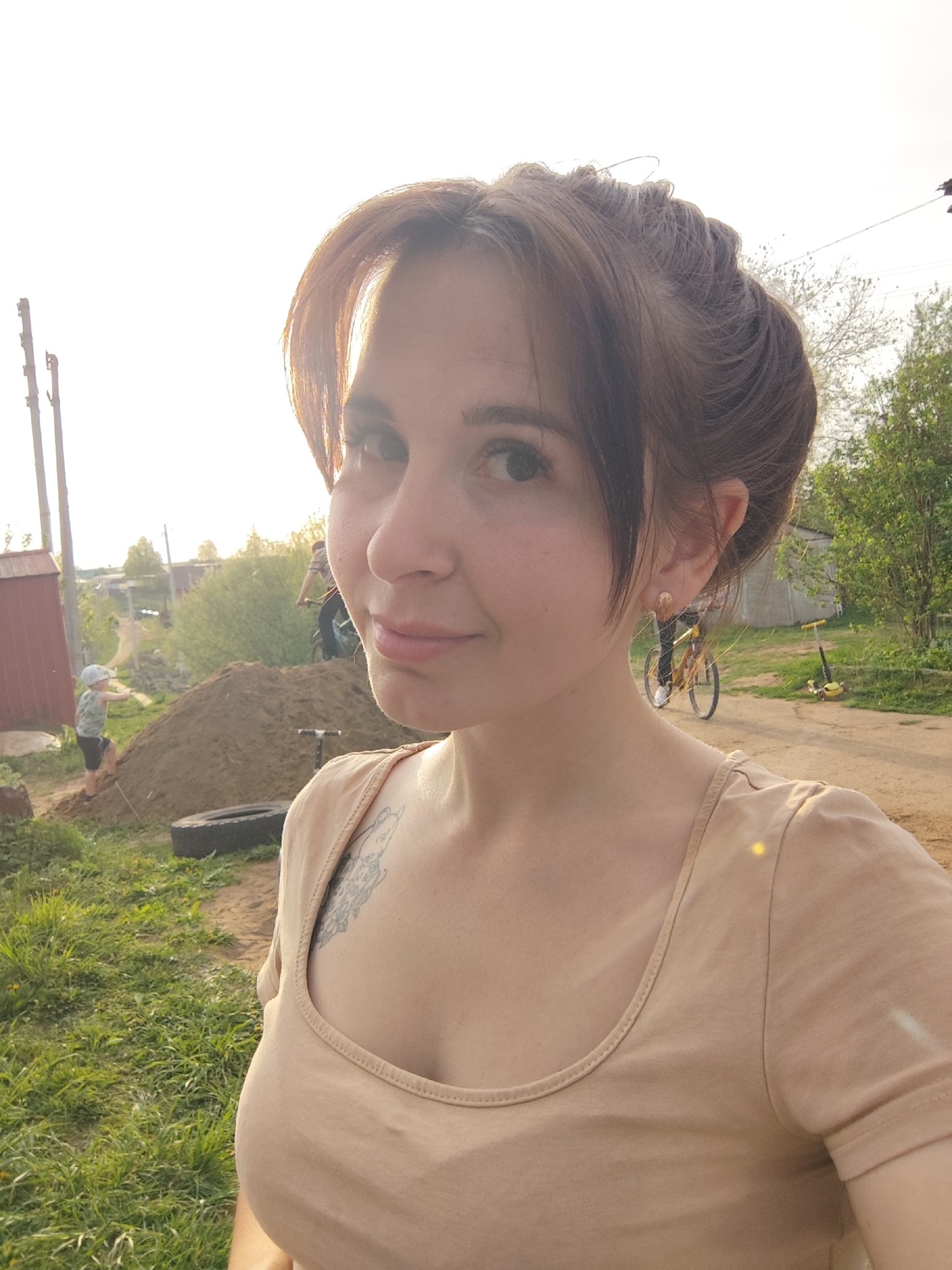 A young woman is taking a selfie outdoors in a rural or suburban setting. She is wearing a beige scoop-neck t-shirt and has her brown hair styled up with bangs. Her face is partially lit by the sun, and she's wearing earrings. A tattoo is visible on her upper chest.
In the background, a dirt road is visible, with greenery on either side. A person on a bicycle is riding down the road, and a yellow scooter is parked nearby. To the left, a small boy in a cap is standing next to a large pile of dirt, with a tire lying on the ground nearby. A small red building and power lines are also visible in the background.
The location is likely Фоки, Russia, given the prompt's specifications.